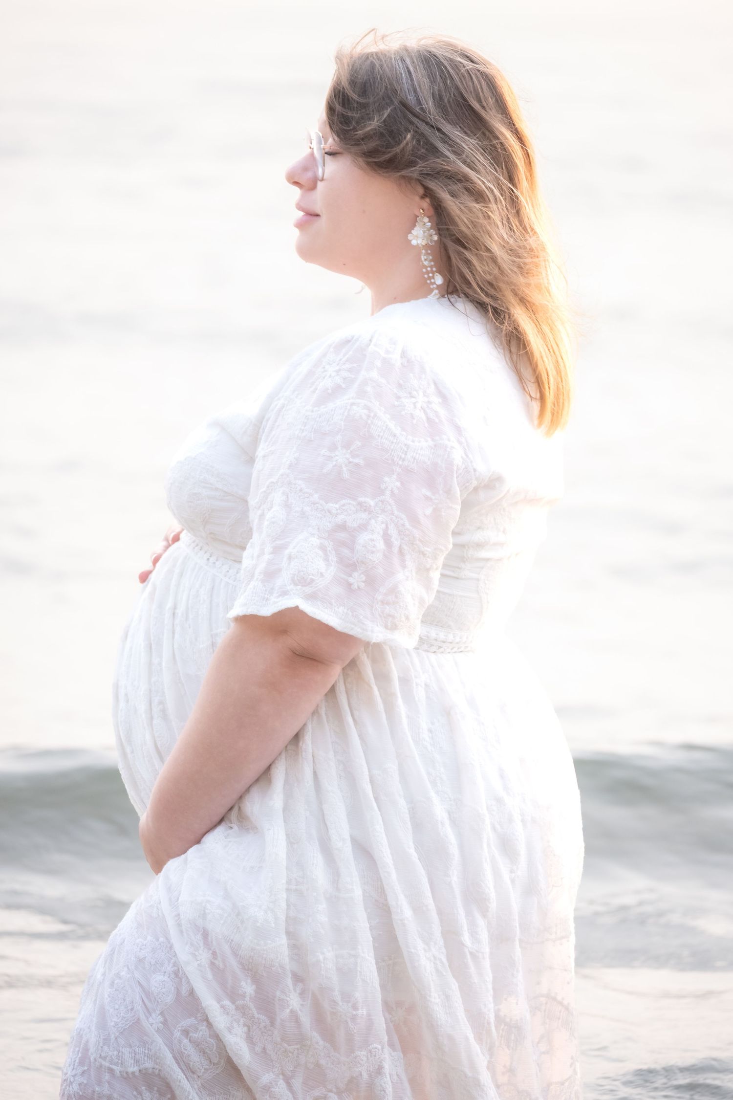 Pregnant woman in white lace dress cradling baby bump by the ocean in soft golden light.