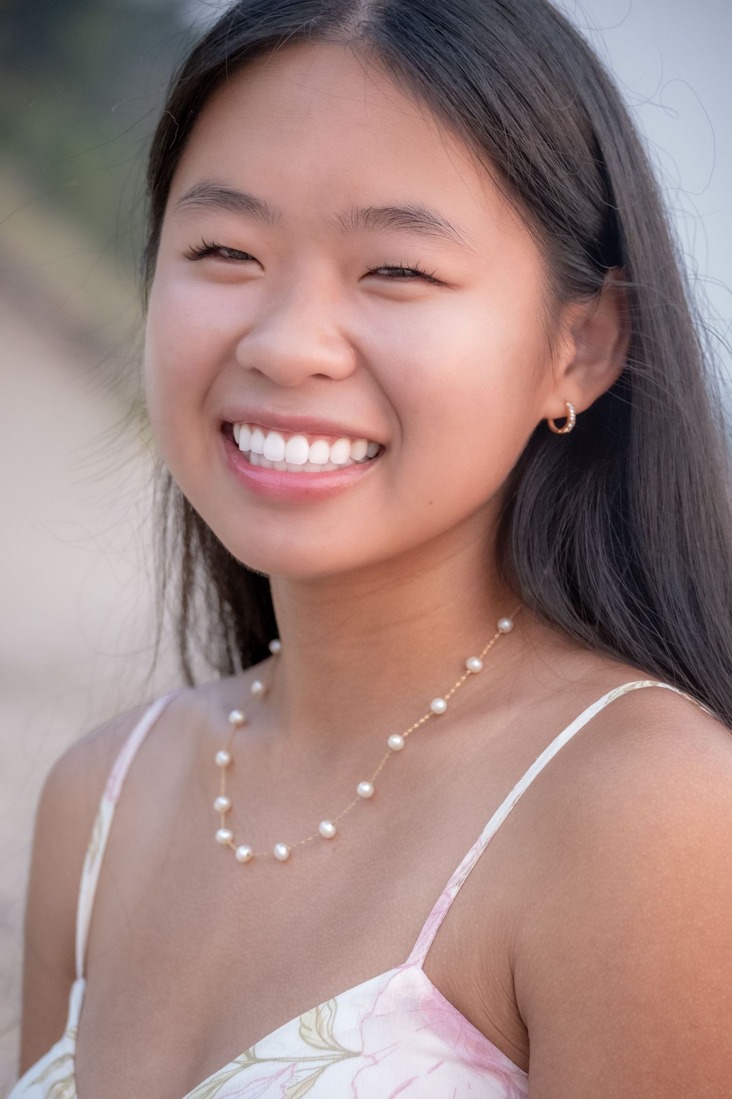 Smiling young Asian woman wearing a pearl necklace and floral dress, photographed outdoors in natural light.