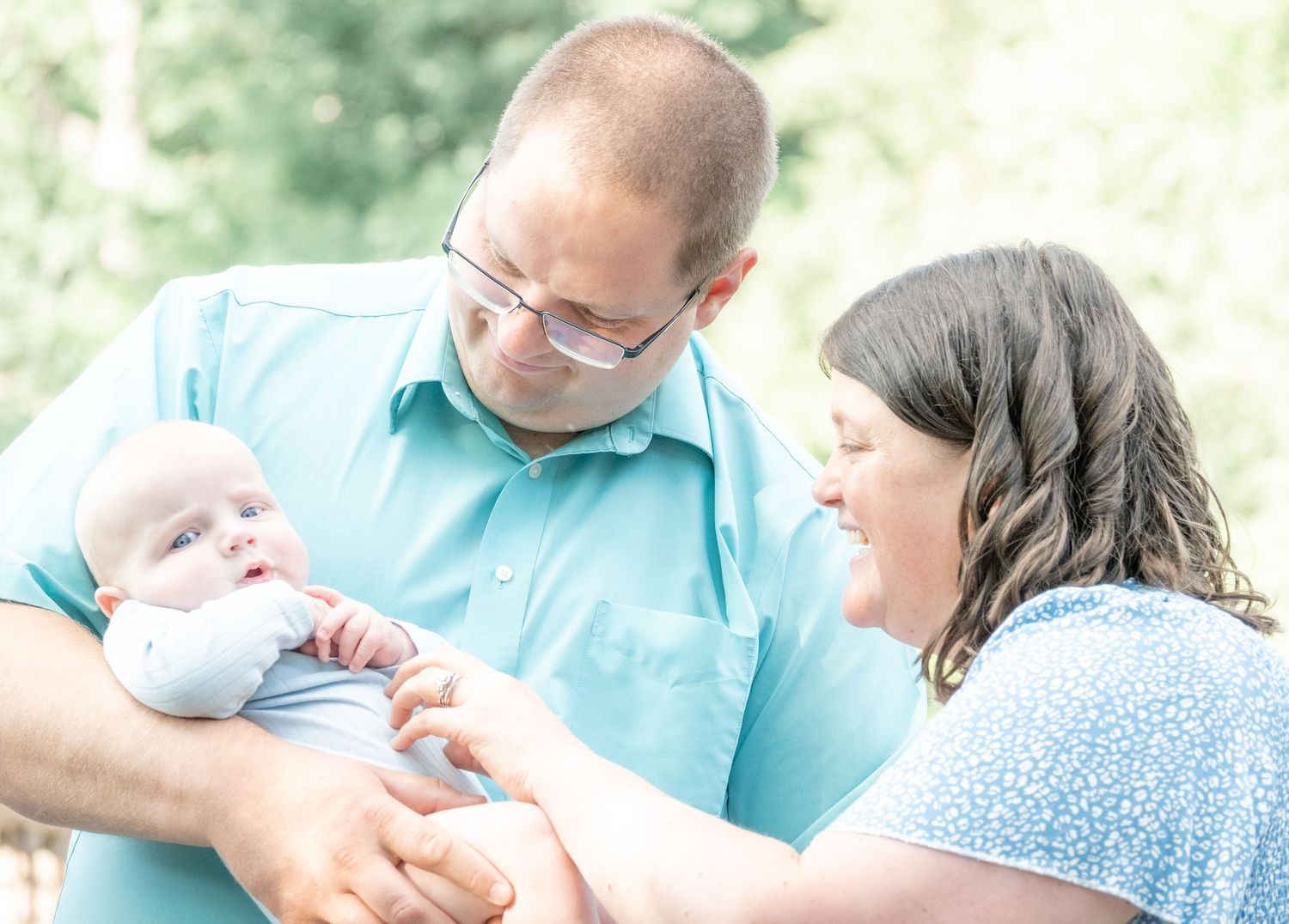 A couple in light blue outfits lovingly gazes at their newborn baby outdoors in a bright, natural setting.