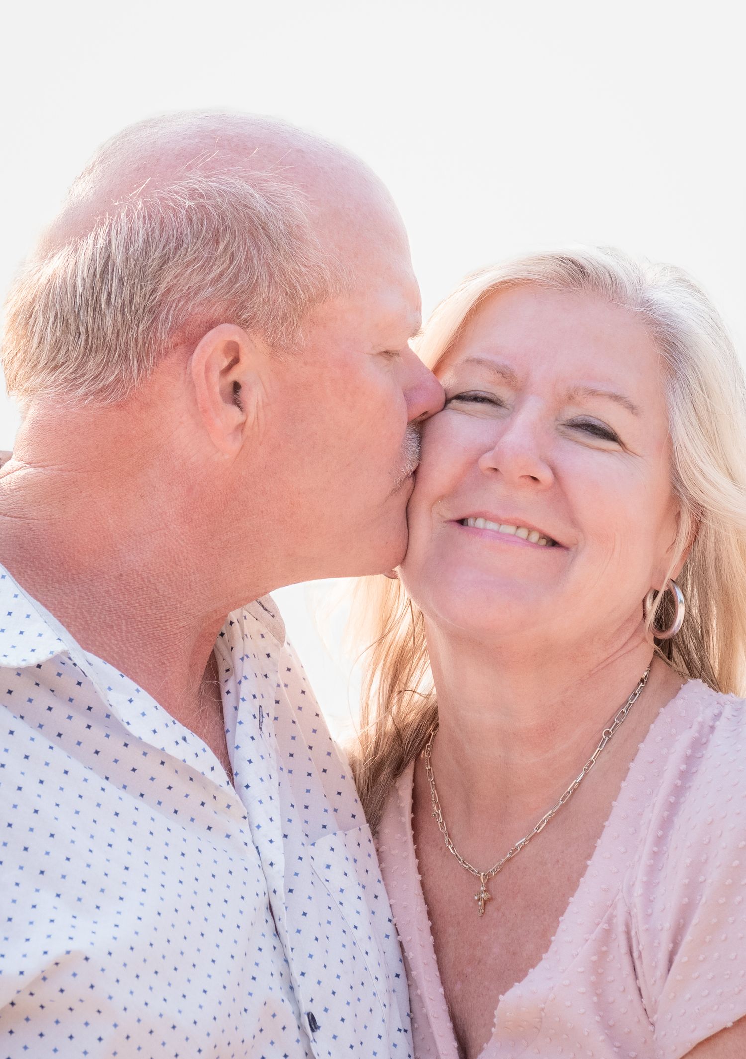 An older couple sharing a tender moment as a man kisses a smiling blonde woman on the cheek outdoors.