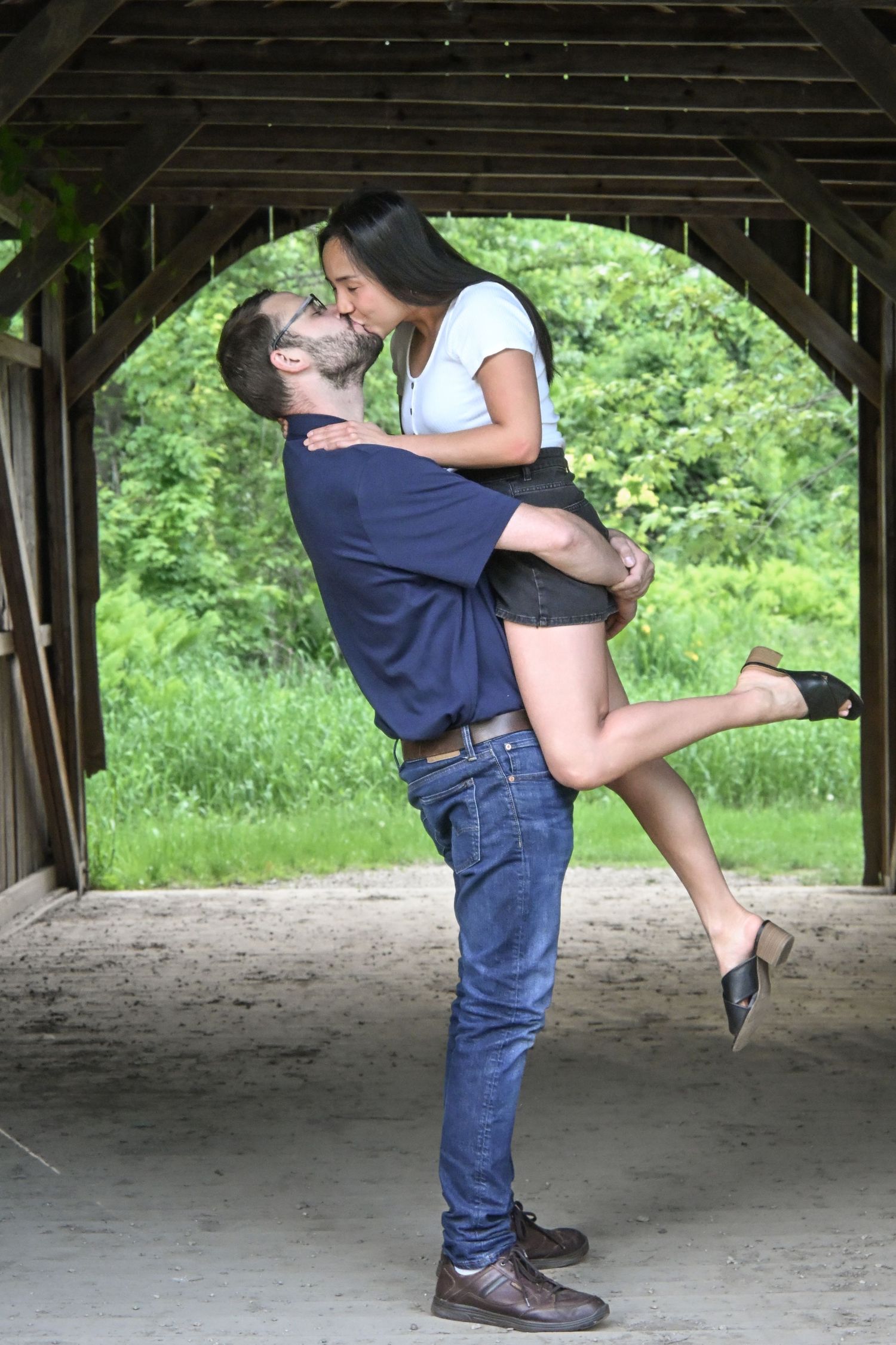 Man lifting woman for a kiss under a rustic covered wooden structure with green trees in the background.