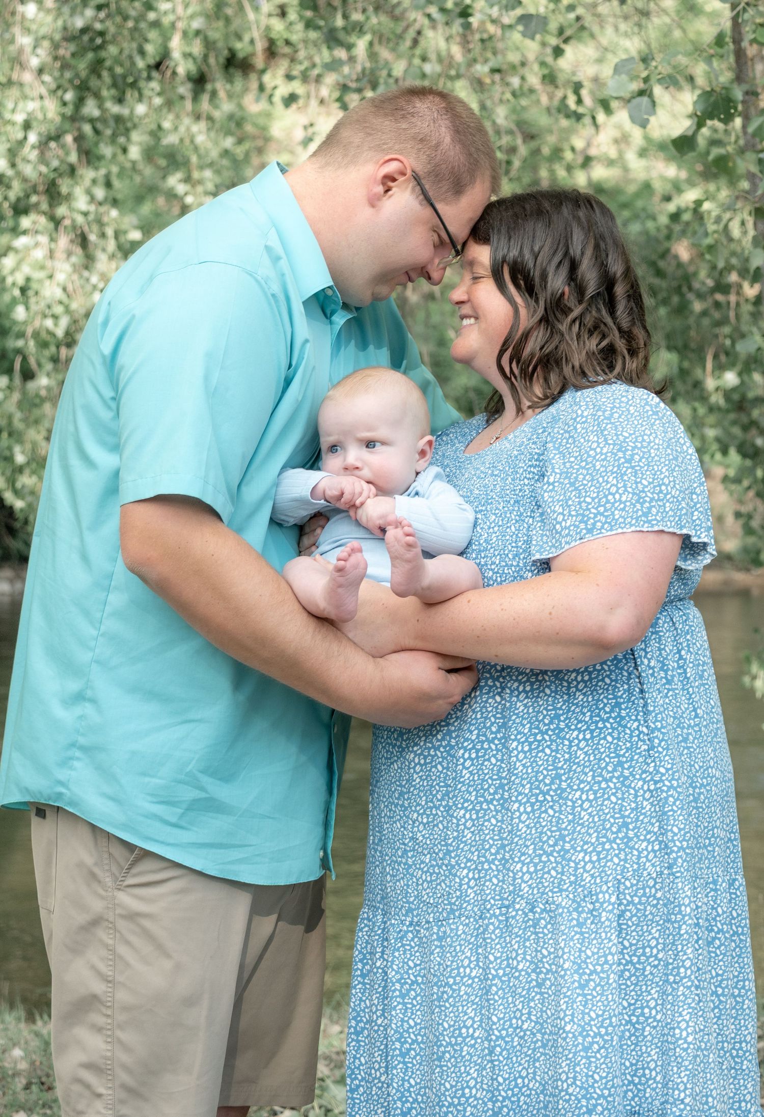 A young couple in matching blue outfits tenderly hold their smiling baby outdoors near lush green trees.