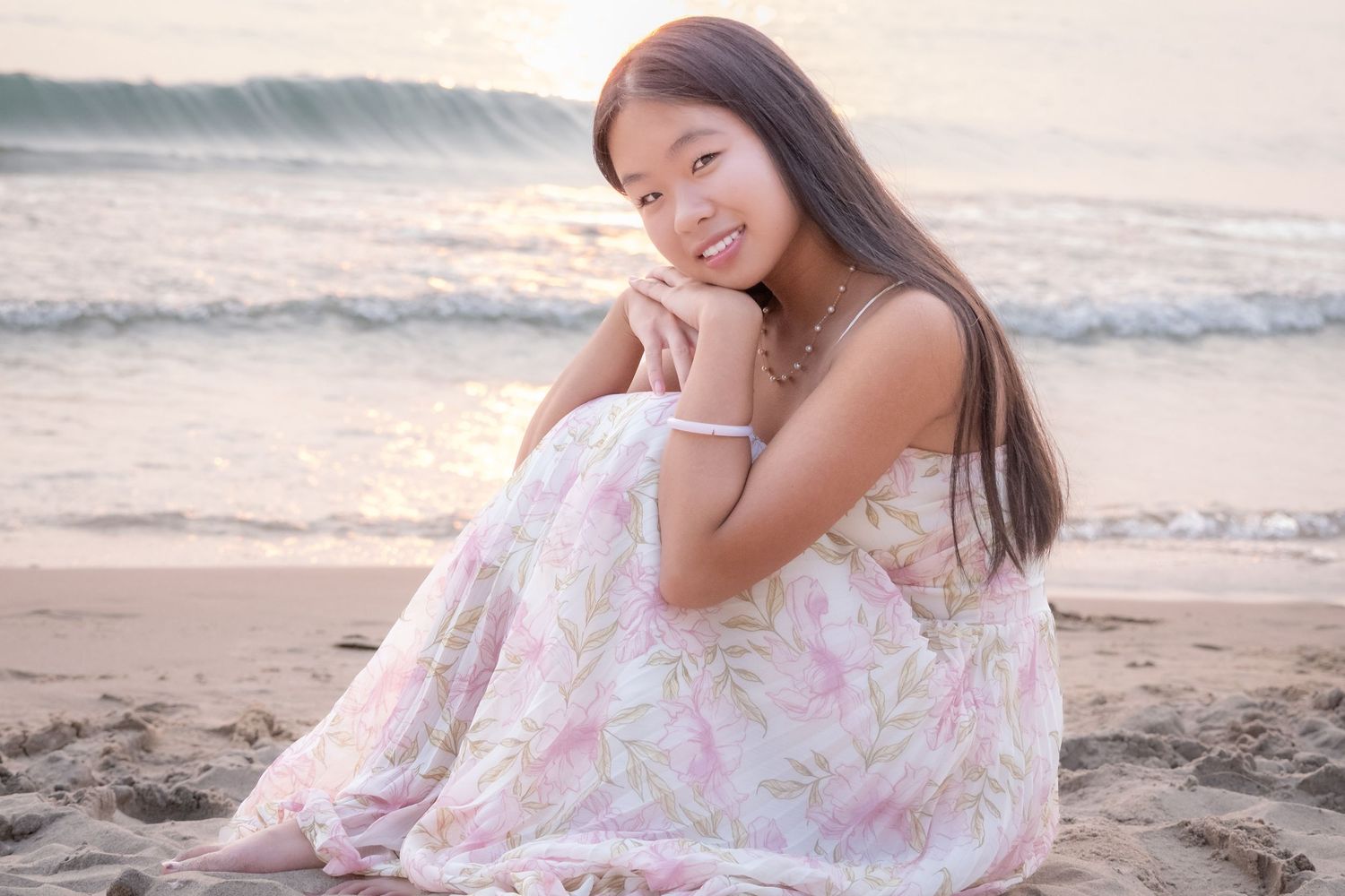 Young Asian woman in floral dress sitting on sandy beach, smiling warmly with ocean waves in background at sunset.