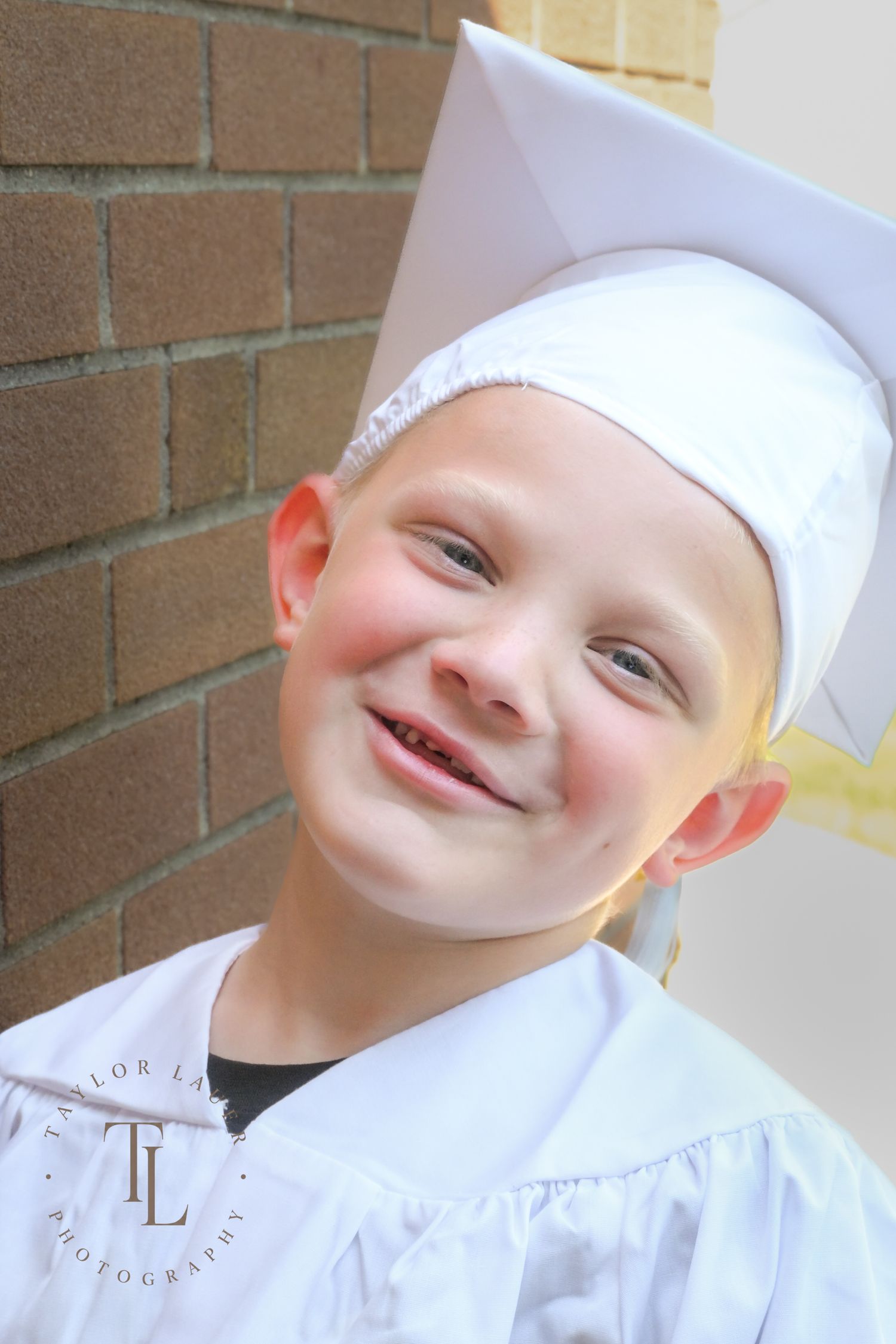 Cheerful young boy in white graduation cap and gown smiling against a brick wall background.