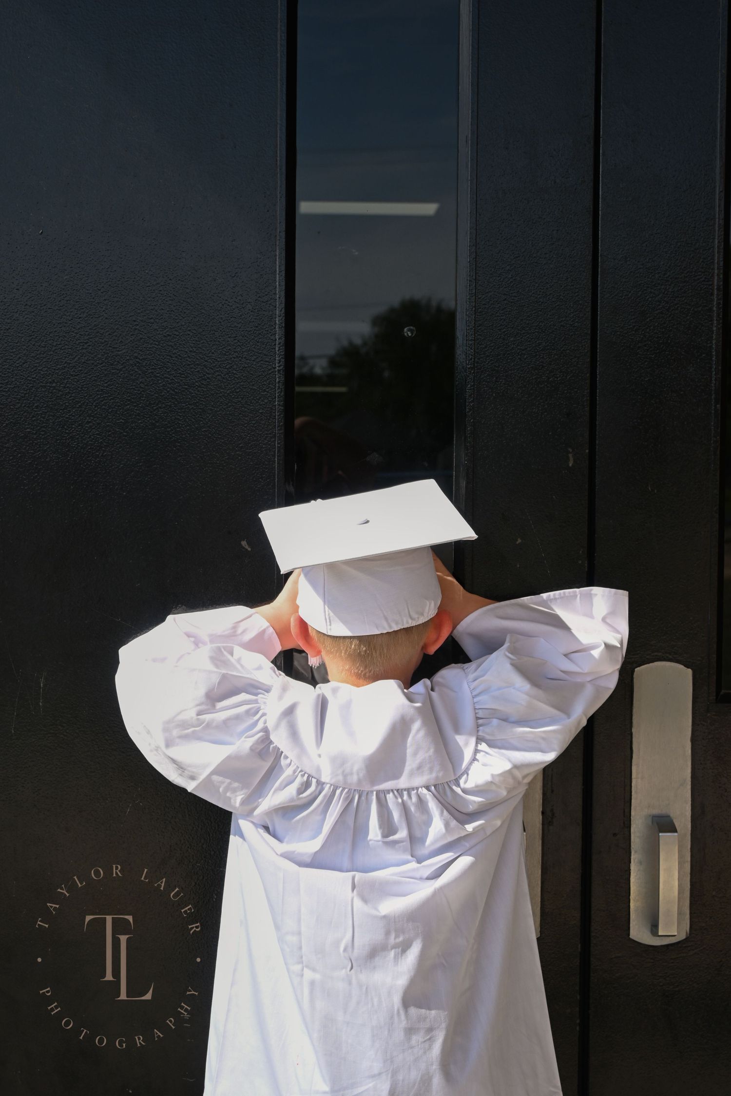 Child in white graduation cap and gown standing with back turned against black double doors.