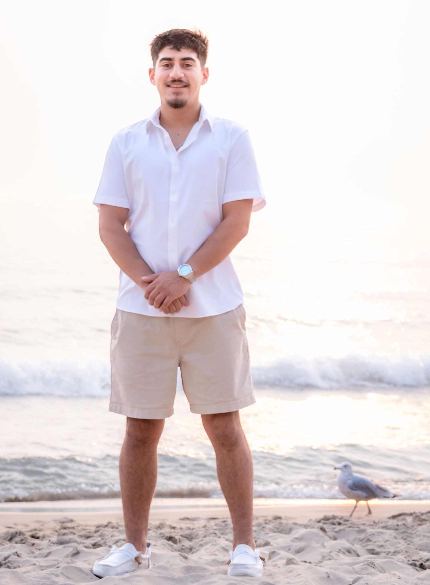Young man in white shirt and khaki shorts smiling on sandy beach with seagull nearby at sunset.