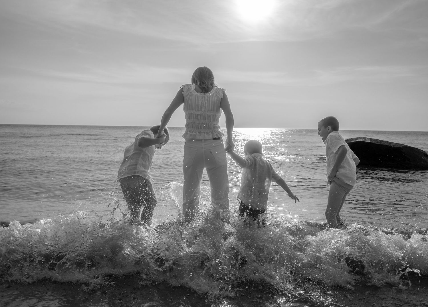 Black and white photo of a parent holding hands with three children playing in ocean waves on a sunny beach.