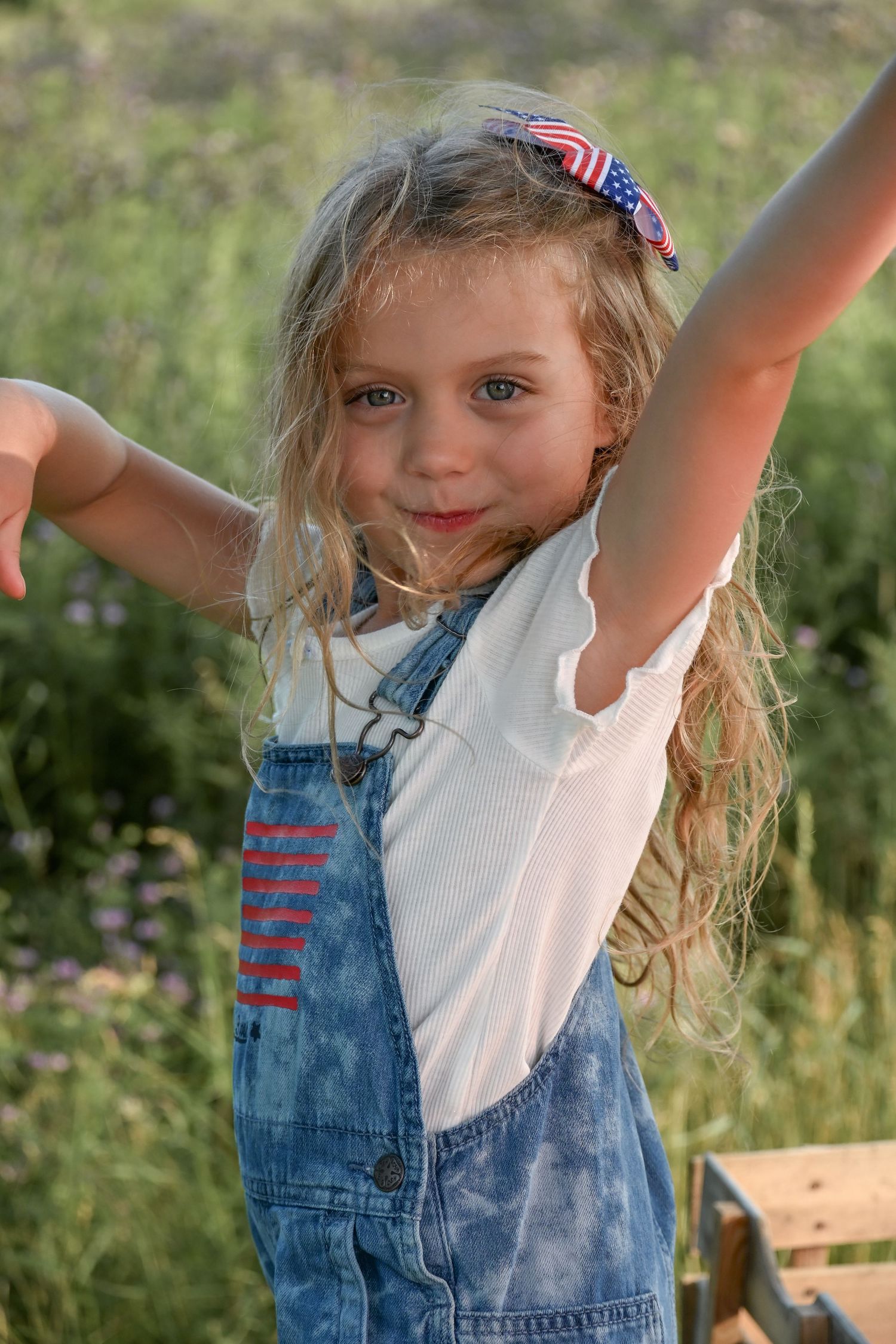 Happy young girl in denim overalls and American flag bow raises her arms outdoors in a sunny field.