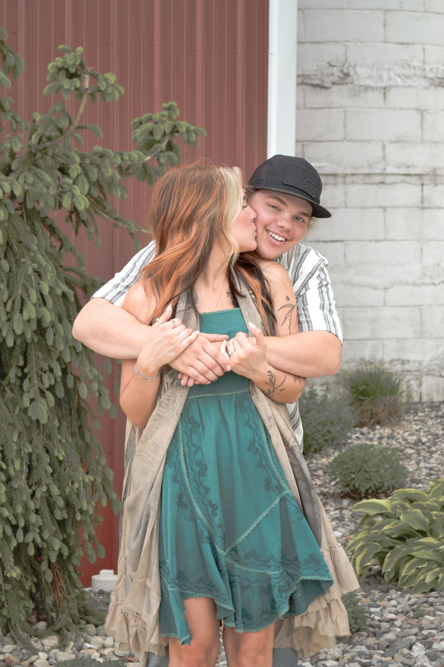 A smiling couple embracing outdoors, the woman kissing the mans cheek near a red barn door.