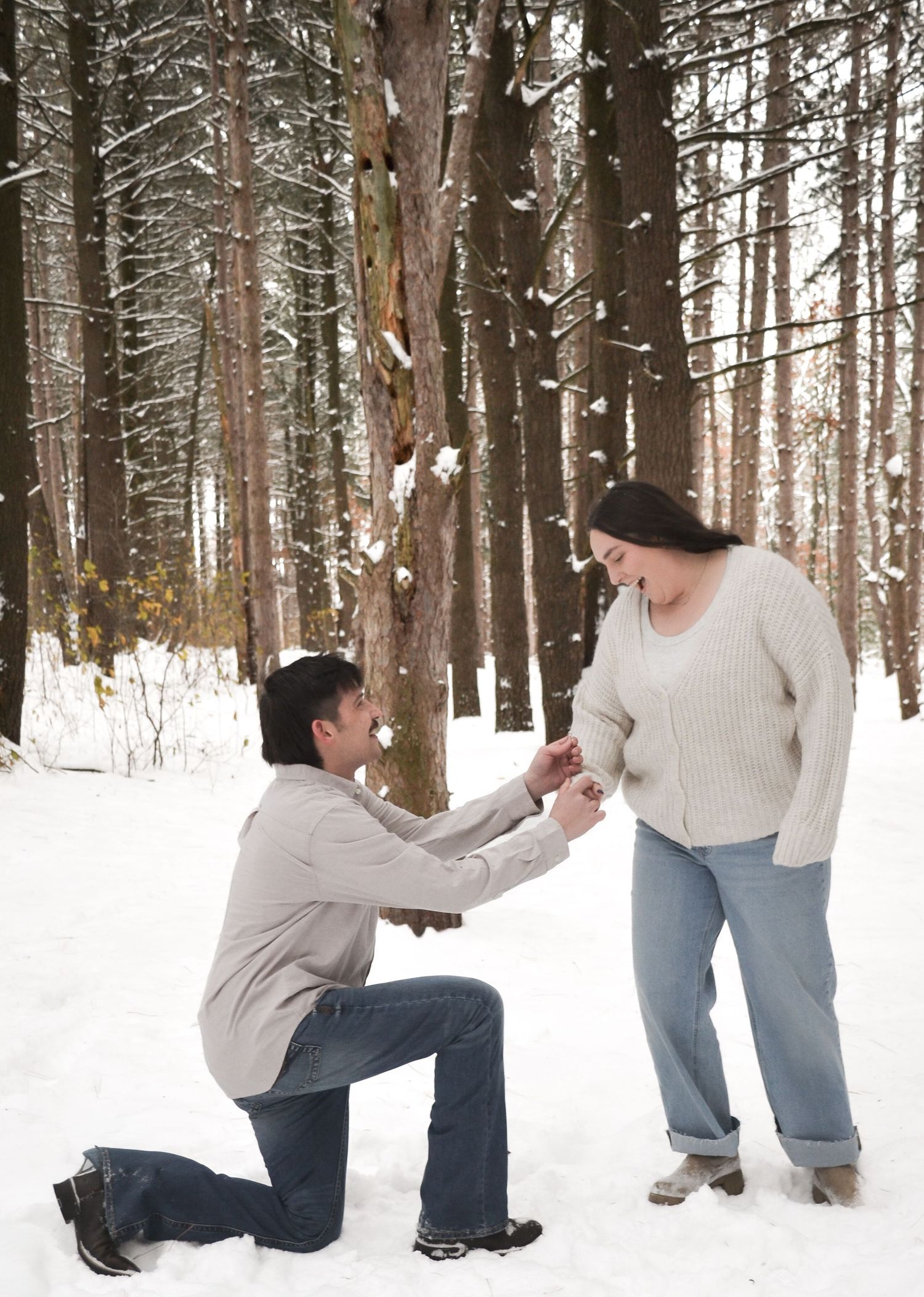 Man proposing on one knee to woman in a snowy winter forest, holding her hands.