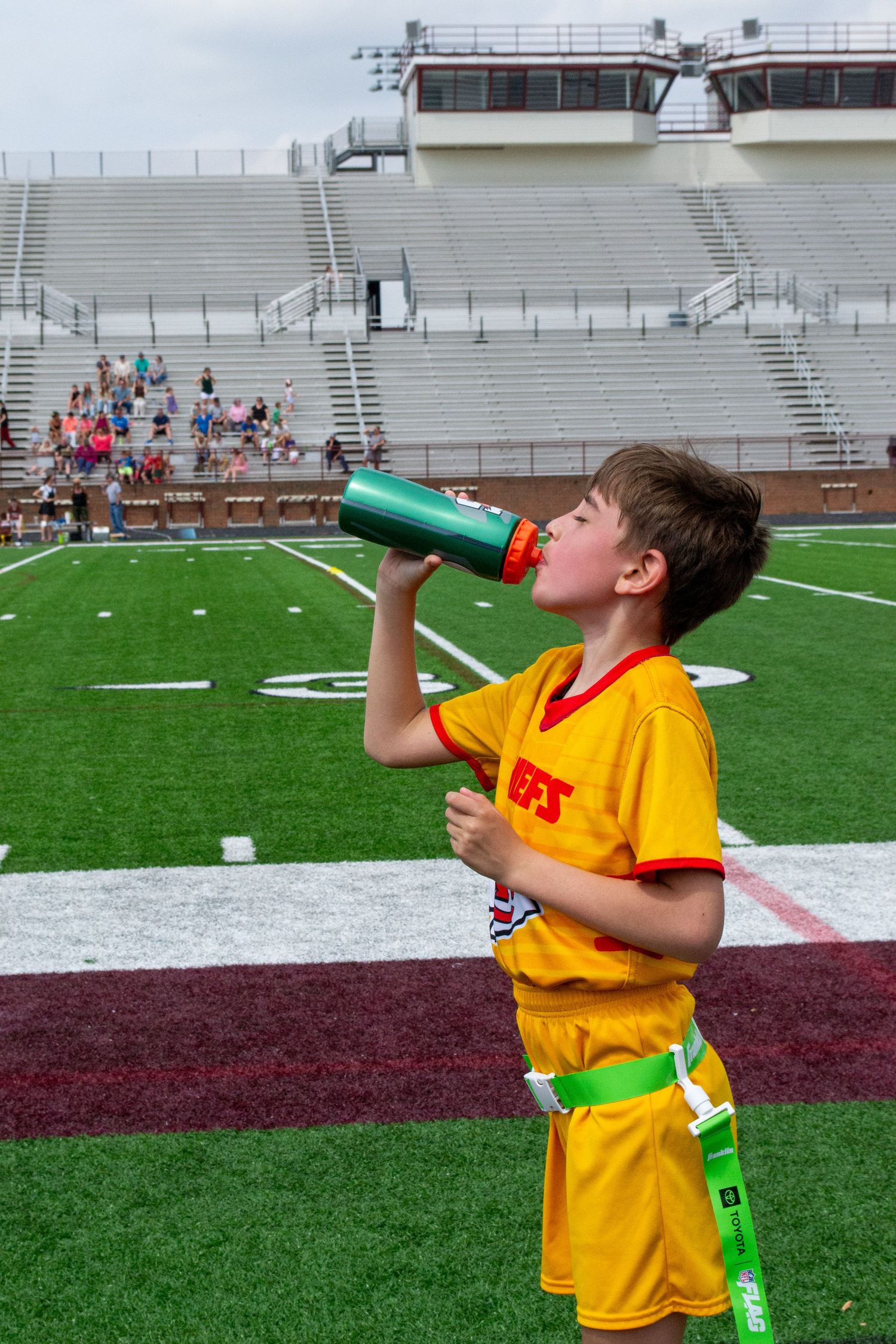 Young boy in yellow Chiefs uniform drinking from water bottle at stadium football field.