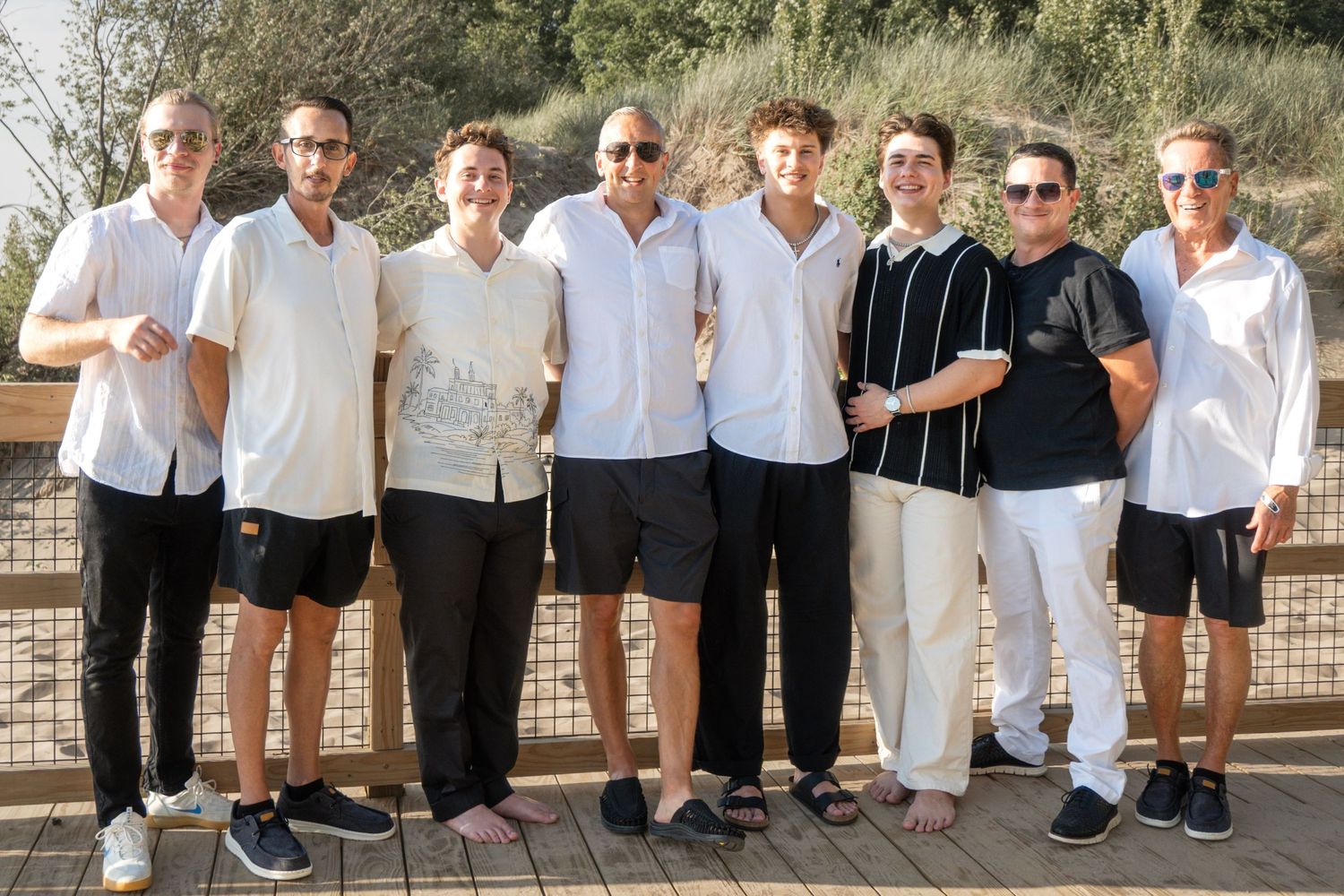 Group of eight men posing together outdoors on a wooden deck, dressed in casual summer outfits of white and black.