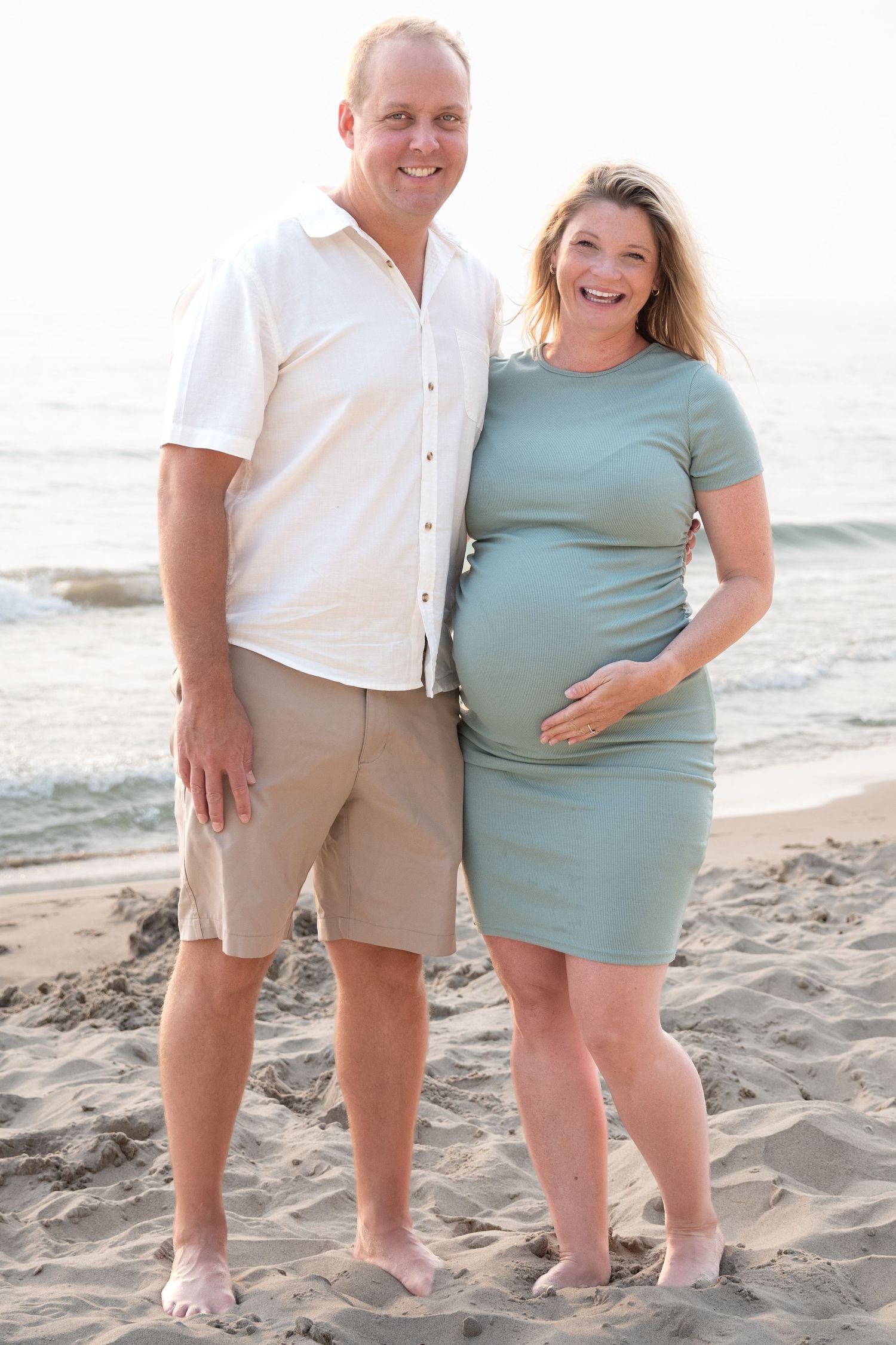 Smiling couple poses on sandy beach, pregnant woman in sage dress cradling belly beside man in white shirt and khaki shorts.