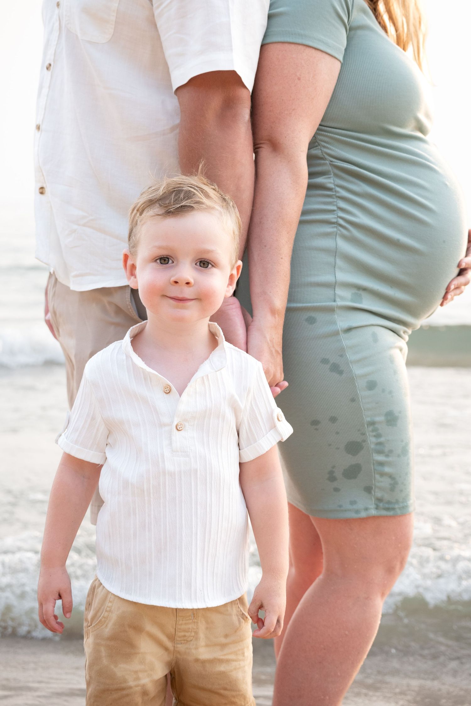 Young boy in white shirt stands before pregnant mother and father on a beach at sunset.