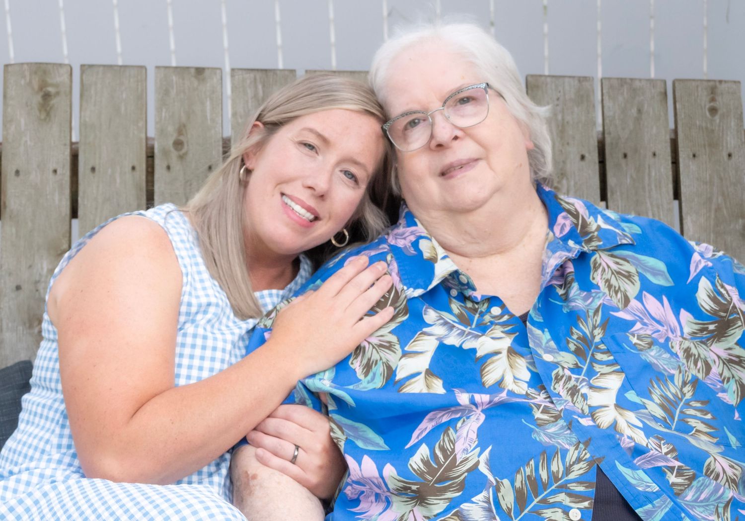 Two smiling women pose together outdoors, one younger in blue dress, one elderly in floral shirt.