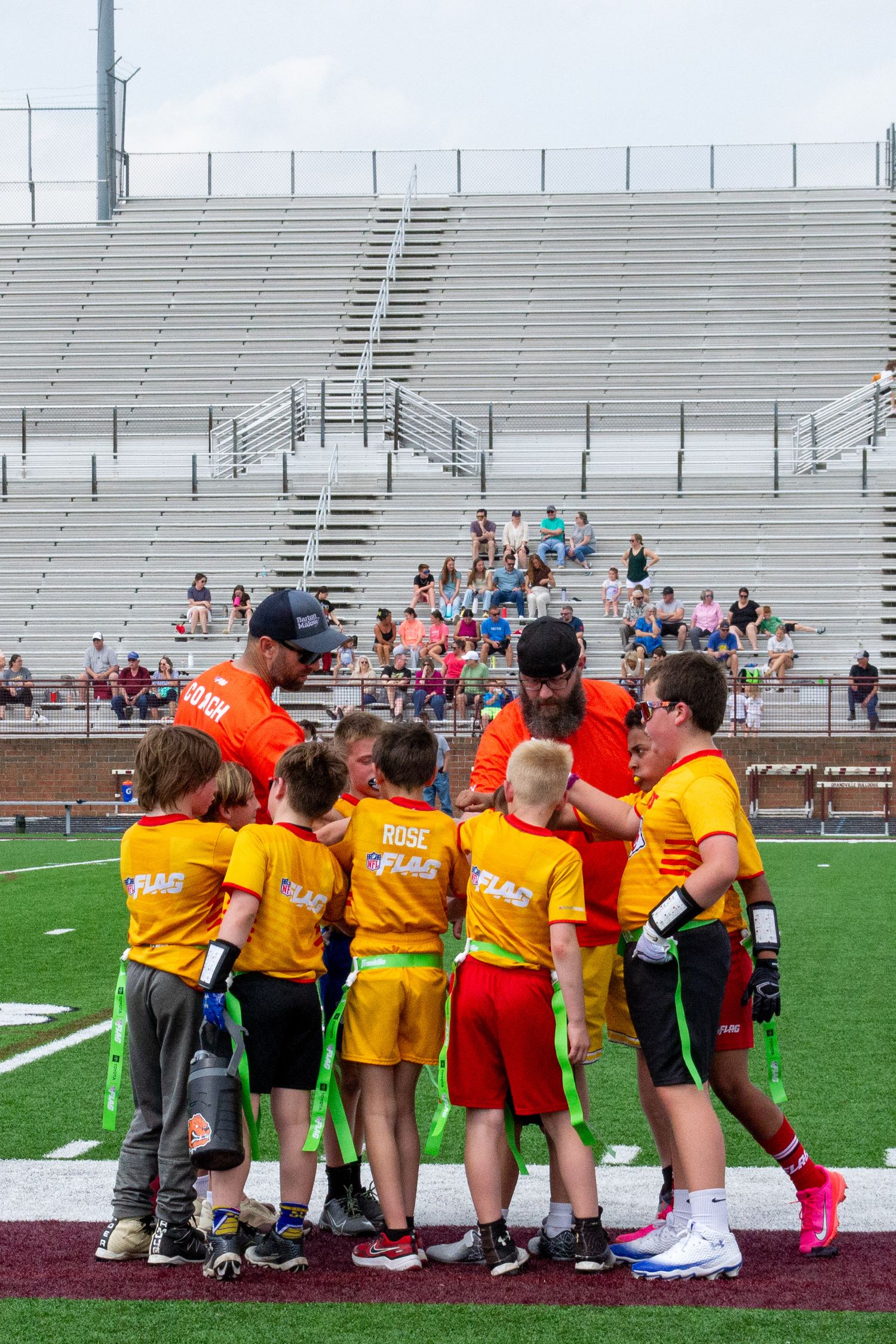 Youth flag football team huddles with coaches in orange jerseys on stadium turf field.