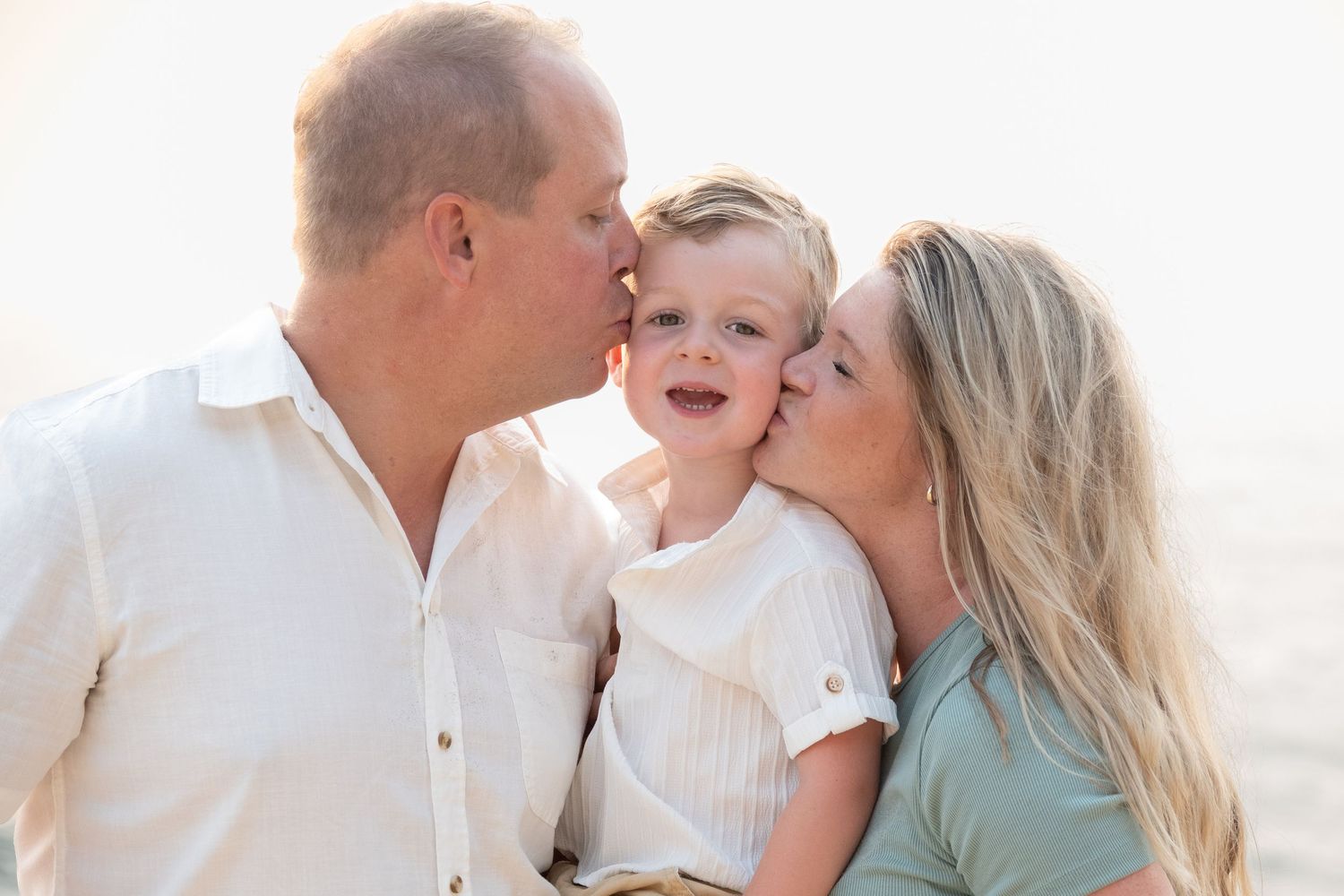 Two parents lovingly kissing their smiling toddler on the cheeks in a bright, warmly lit outdoor family portrait.