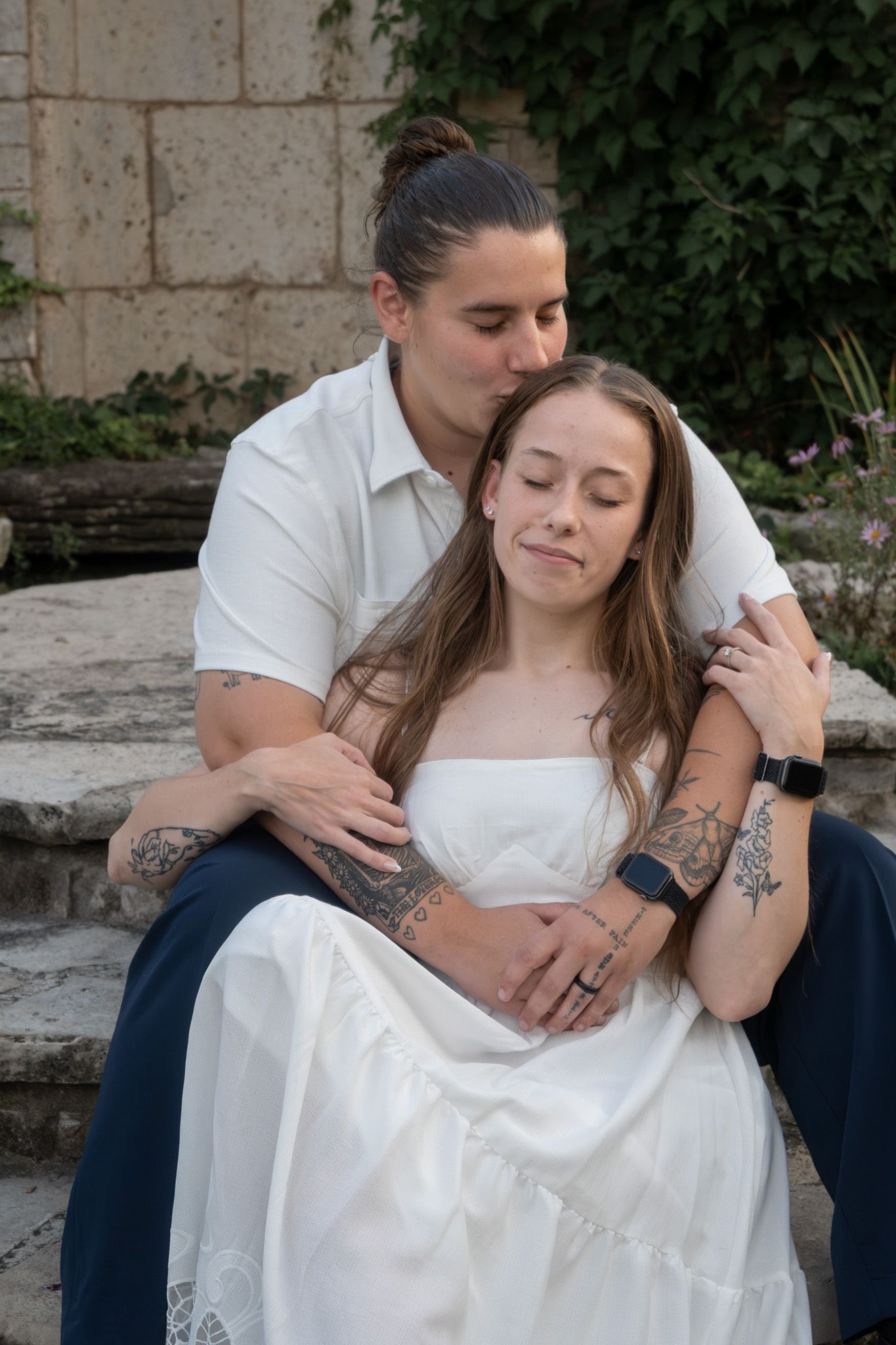 Couple in wedding attire sitting on stone steps, groom kissing bride's head, both with tattoos, outdoors.