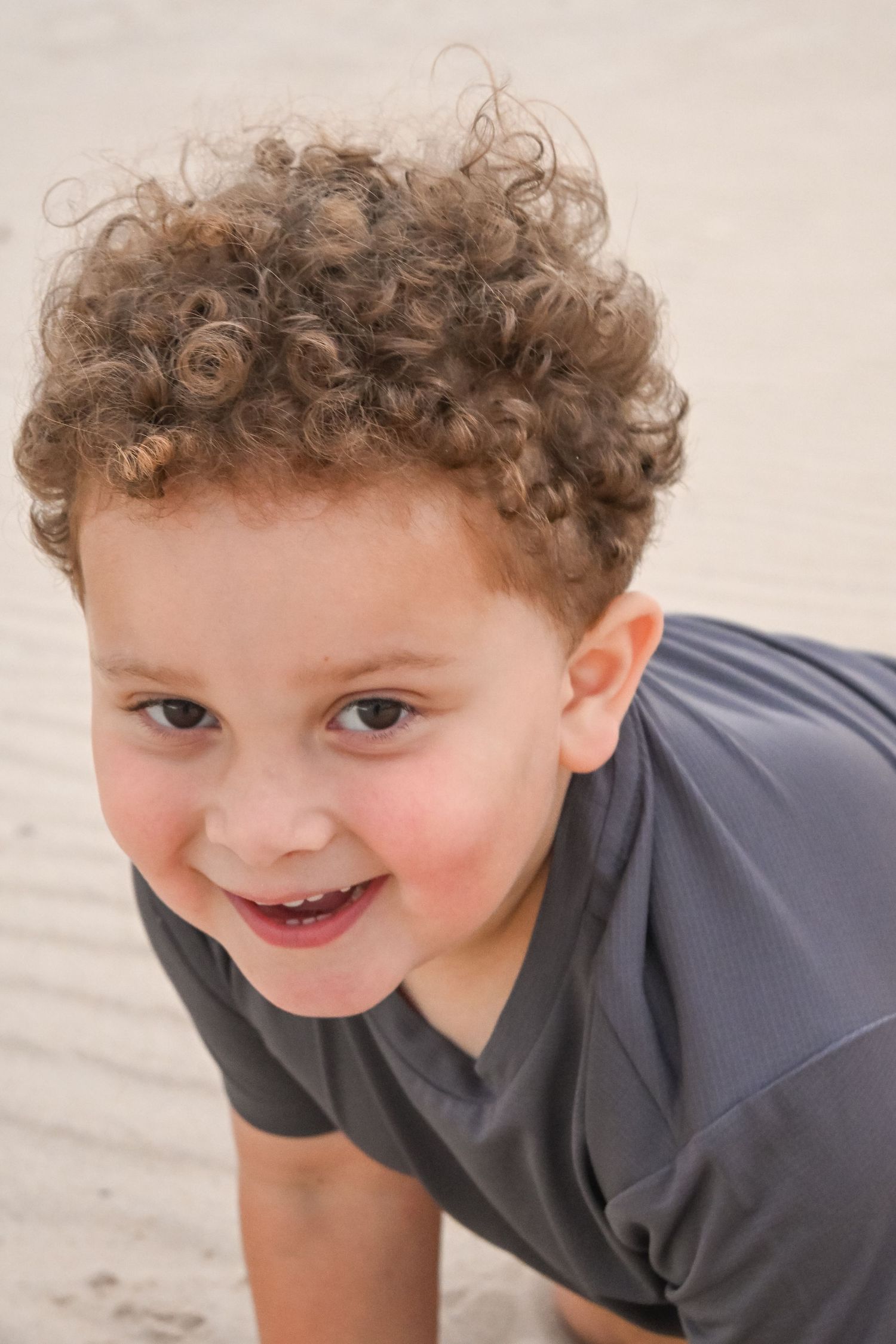 A curly-haired young boy smiles playfully while crouching on sandy beach.