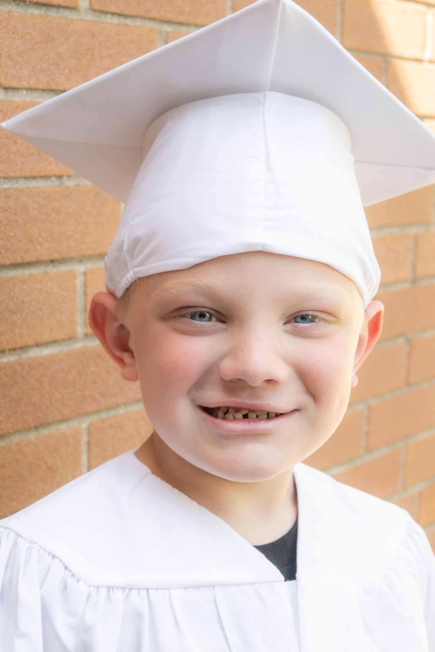 Young child smiling in white graduation cap and gown, standing in front of a brick wall.
