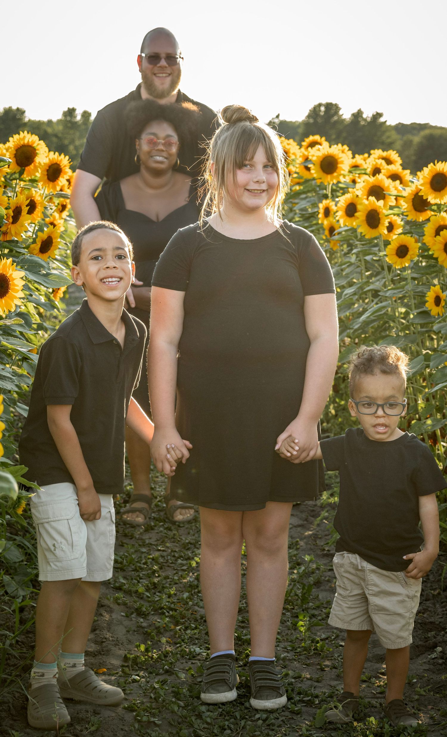 Family of five posing in a sunflower field, wearing matching black outfits, smiling in warm golden hour light.
