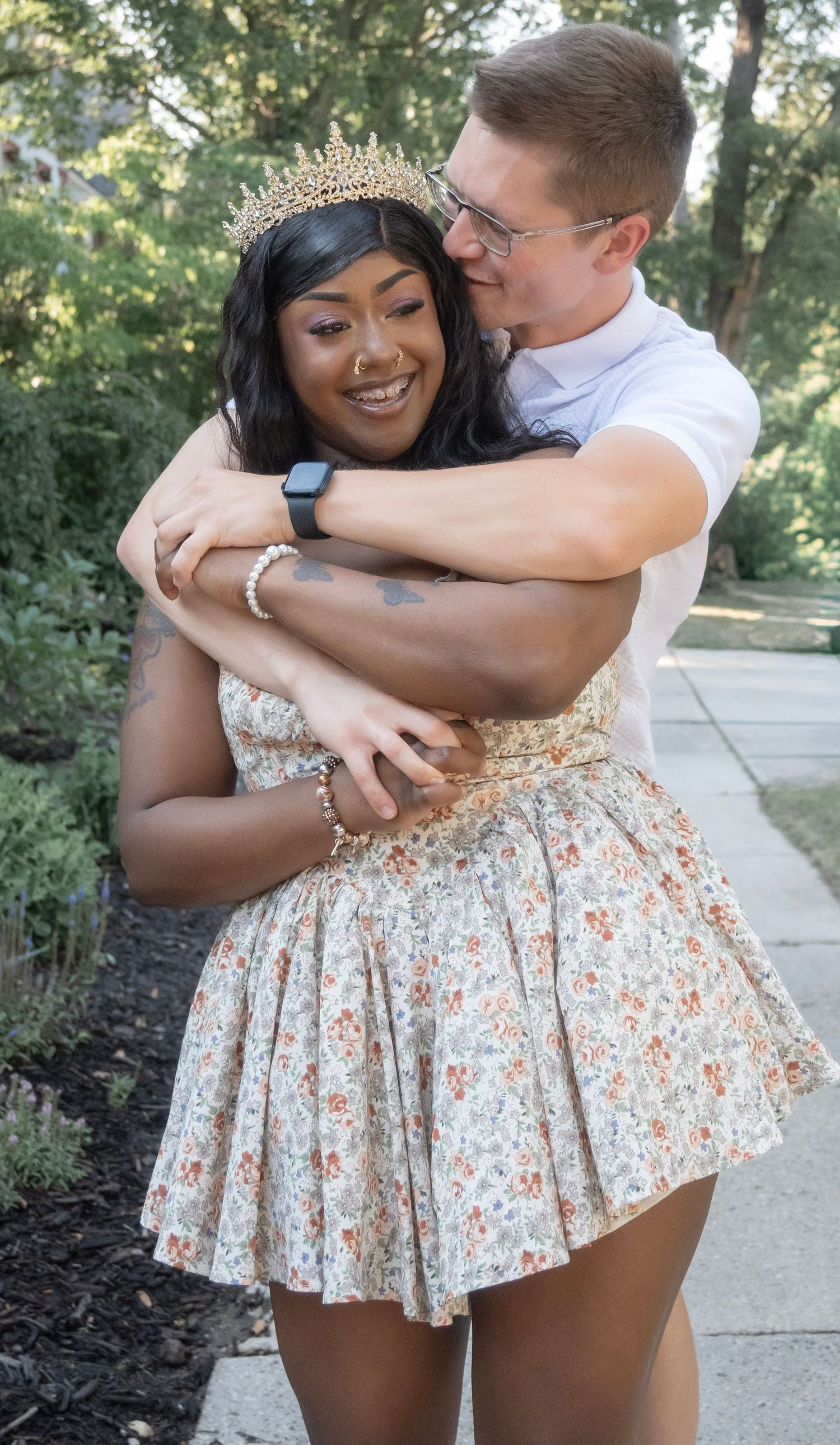 A smiling woman wearing a floral dress and gold crown being embraced by a man outdoors.