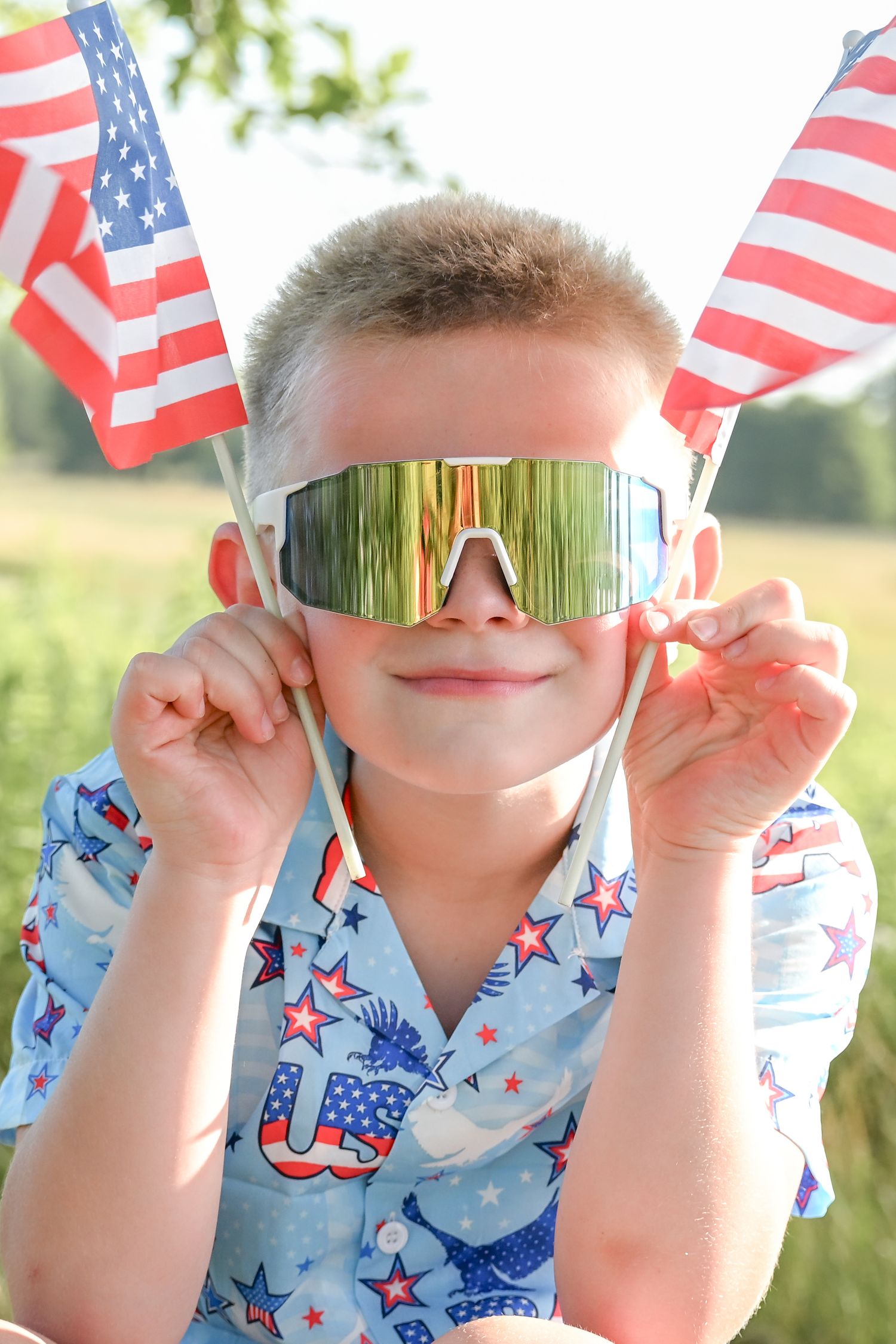 Boy wearing mirrored sunglasses holding two American flags outdoors, dressed in patriotic USA shirt for Fourth of July.