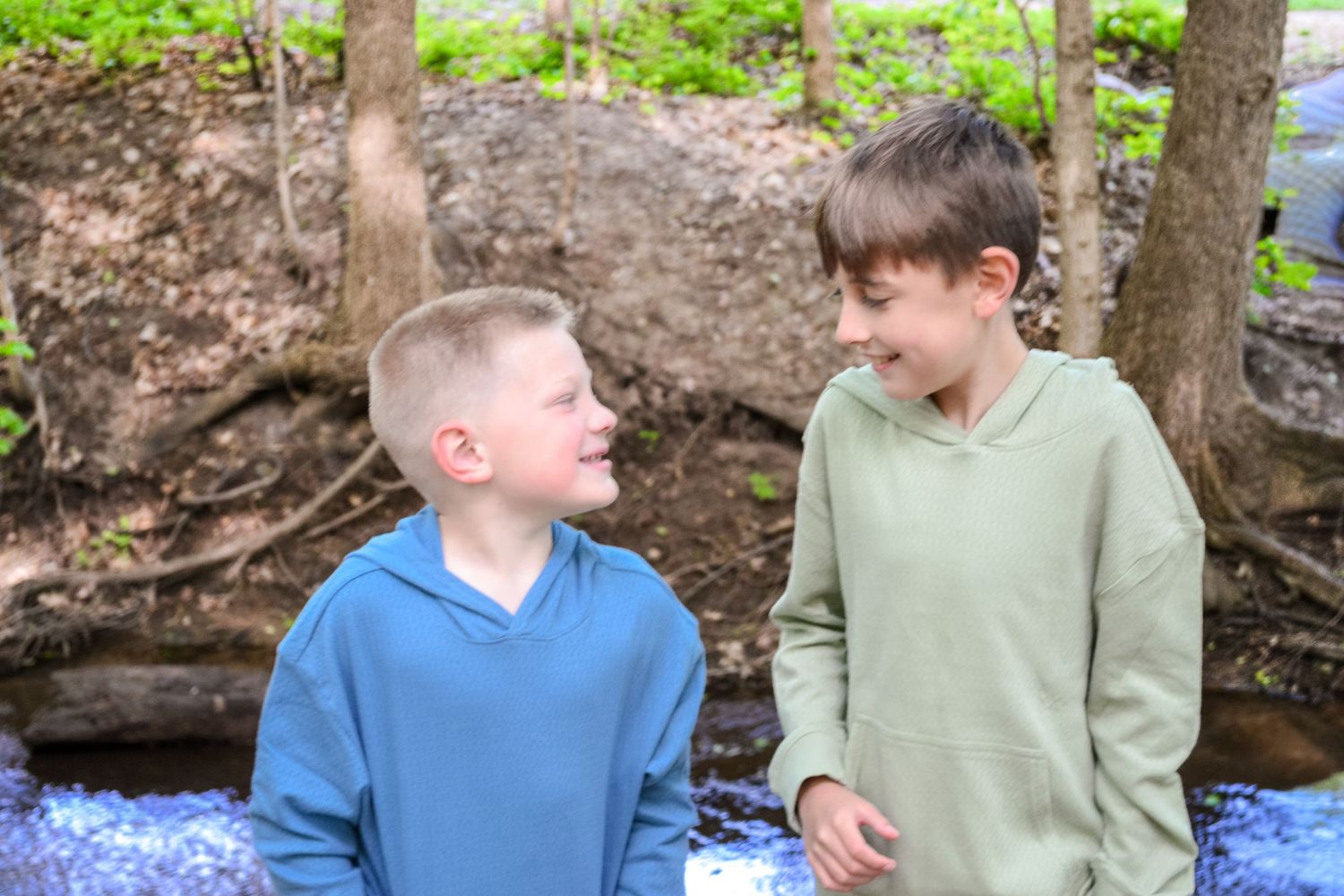 Two boys smiling at each other near a rocky stream in a wooded outdoor setting.