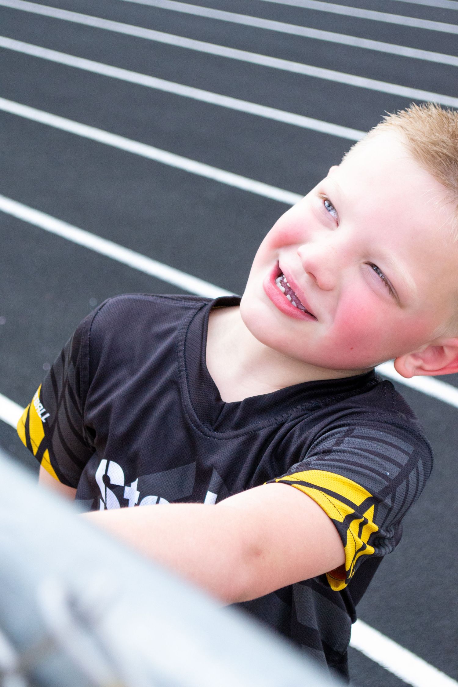 Smiling young boy in black and gold sports jersey leaning on a railing near a track.
