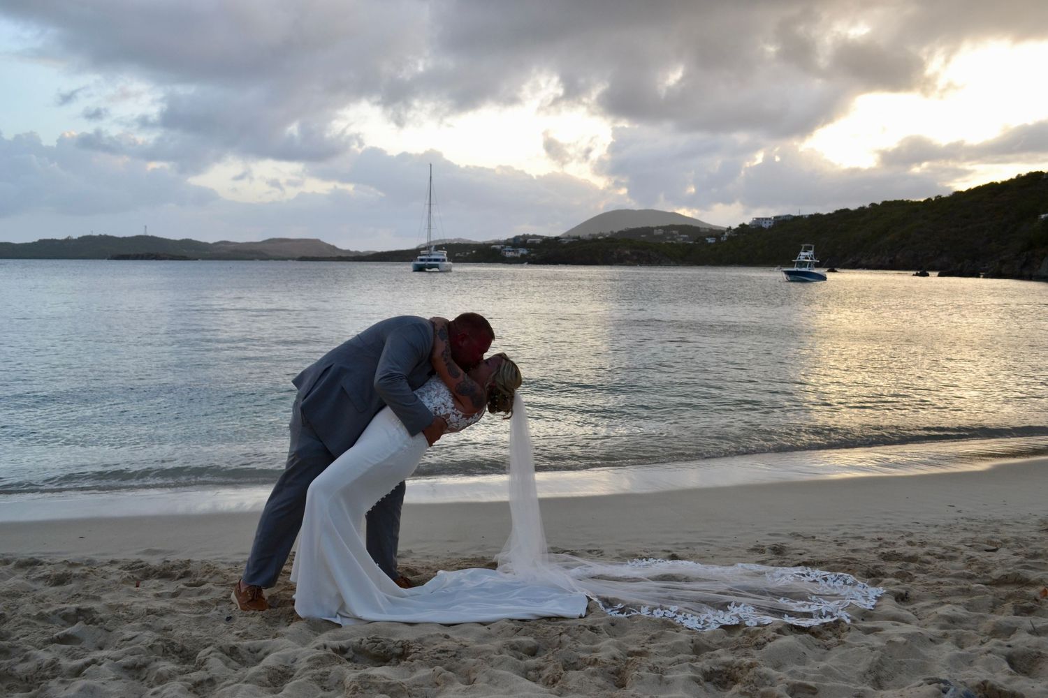 Couple sharing a romantic dip kiss on a sandy beach at sunset with sailboats in the background.