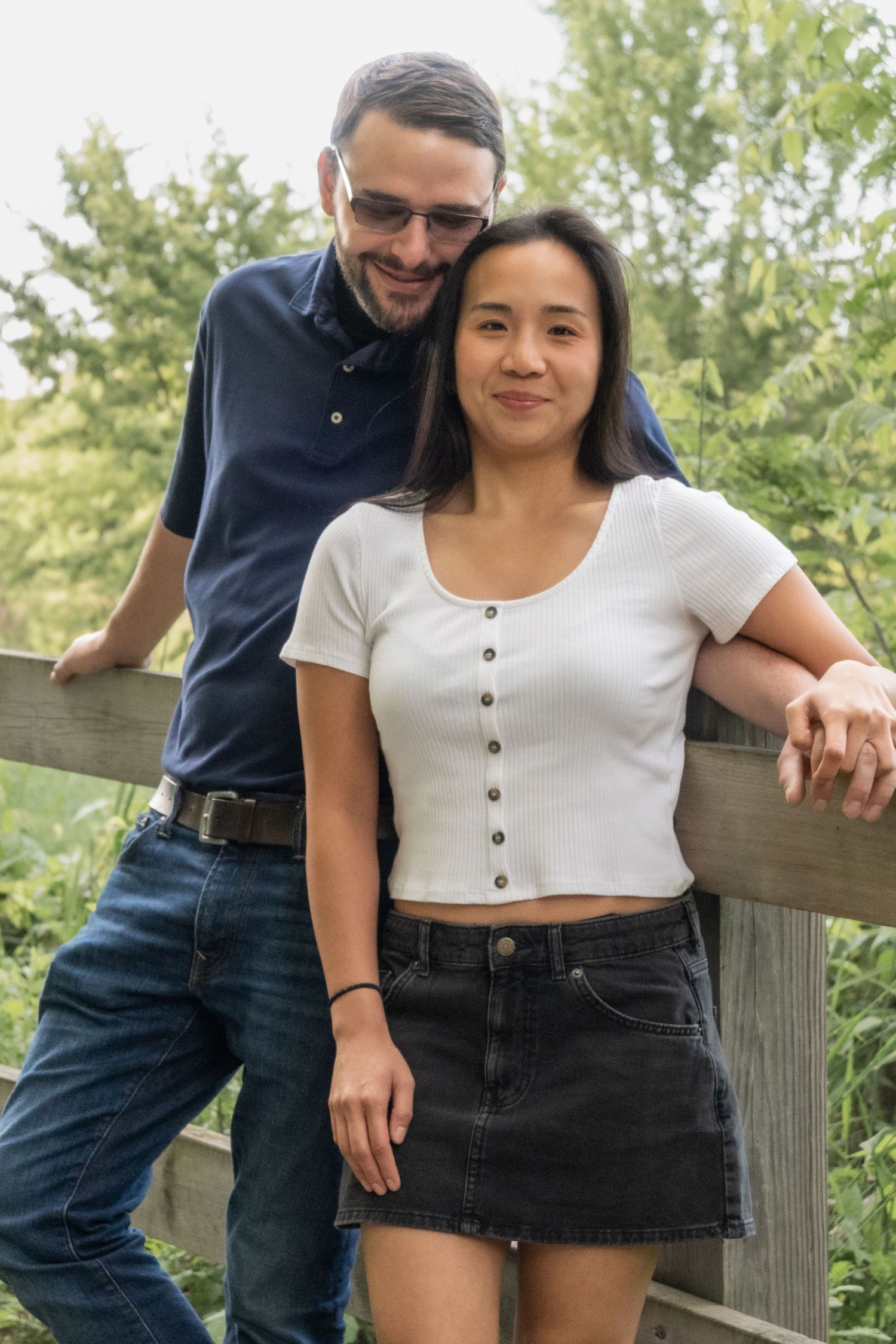 Smiling couple posing by a wooden fence surrounded by lush green trees in an outdoor park setting.