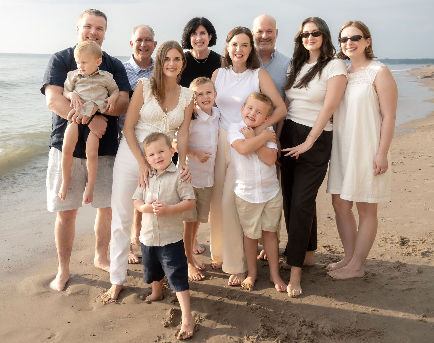 Large family group photo on sandy beach, multi-generational smiling together, wearing white and neutral tones.