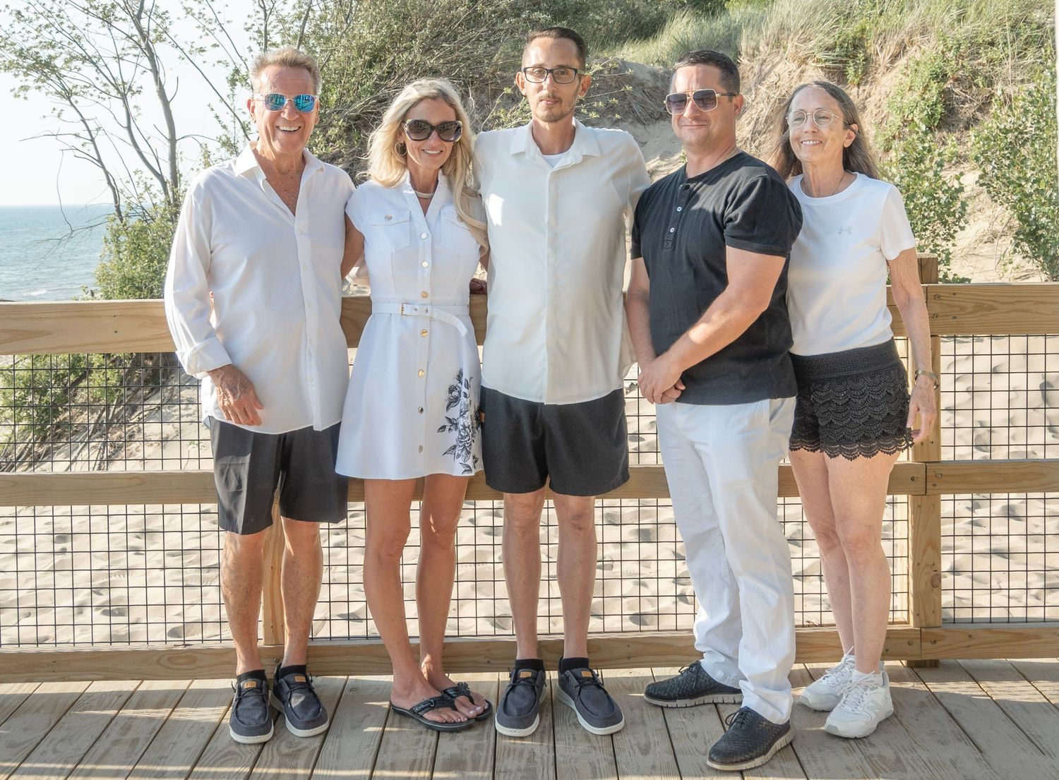 Five adults posing together on a wooden boardwalk near a beach on a sunny day.