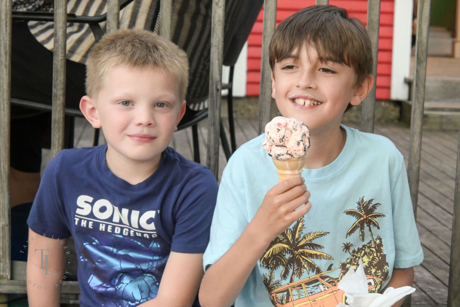 Two young boys smiling while sharing an ice cream cone at an outdoor ice cream shop.