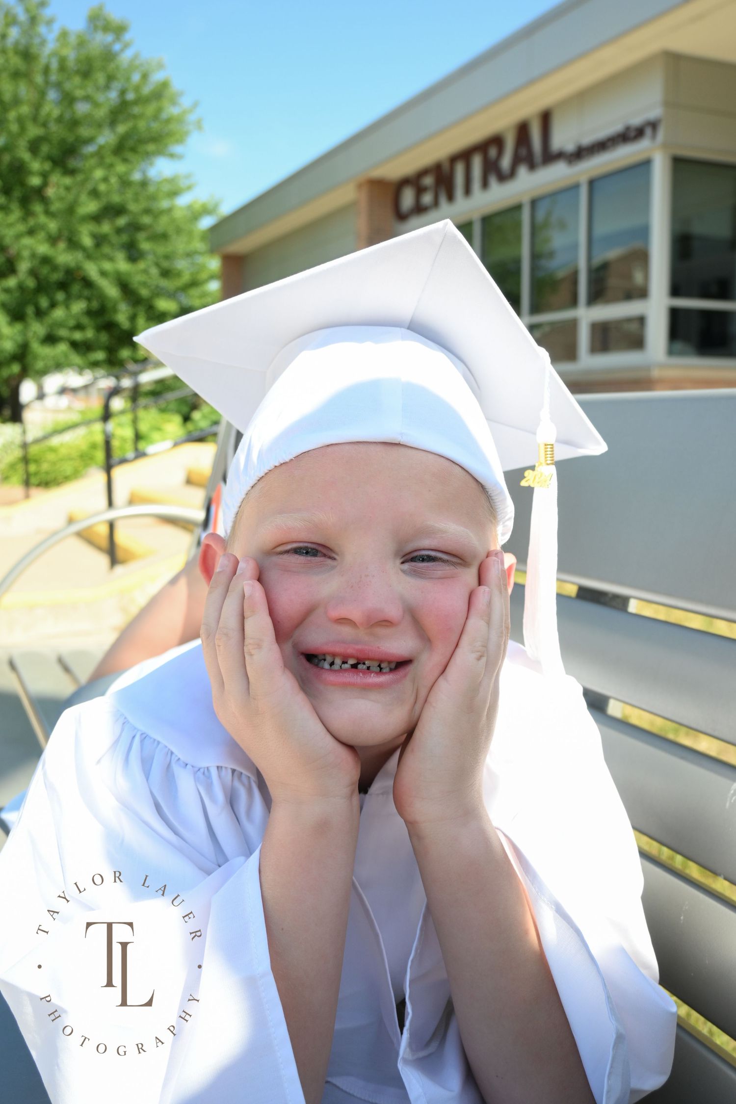 Young child in white kindergarten graduation cap and gown smiling with hands on cheeks outside Central Elementary.