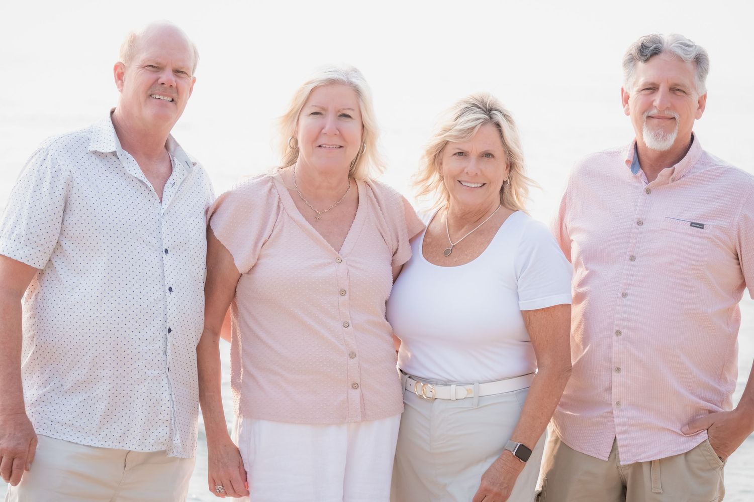 Four smiling adults posing together on a sunny beach, dressed in casual pink and white summer outfits.