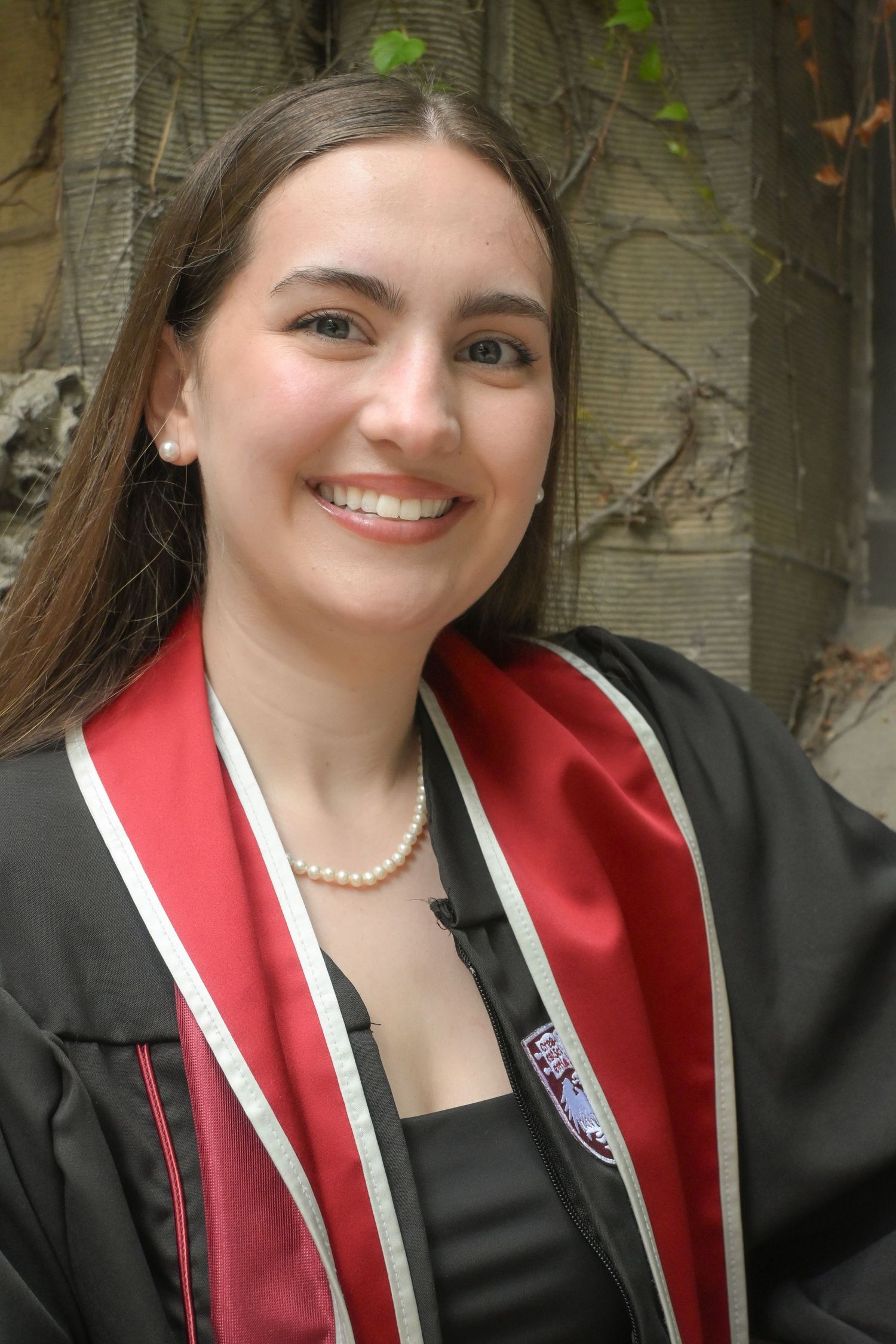 Smiling young woman in black graduation gown with red sash and pearl necklace posing outdoors.
