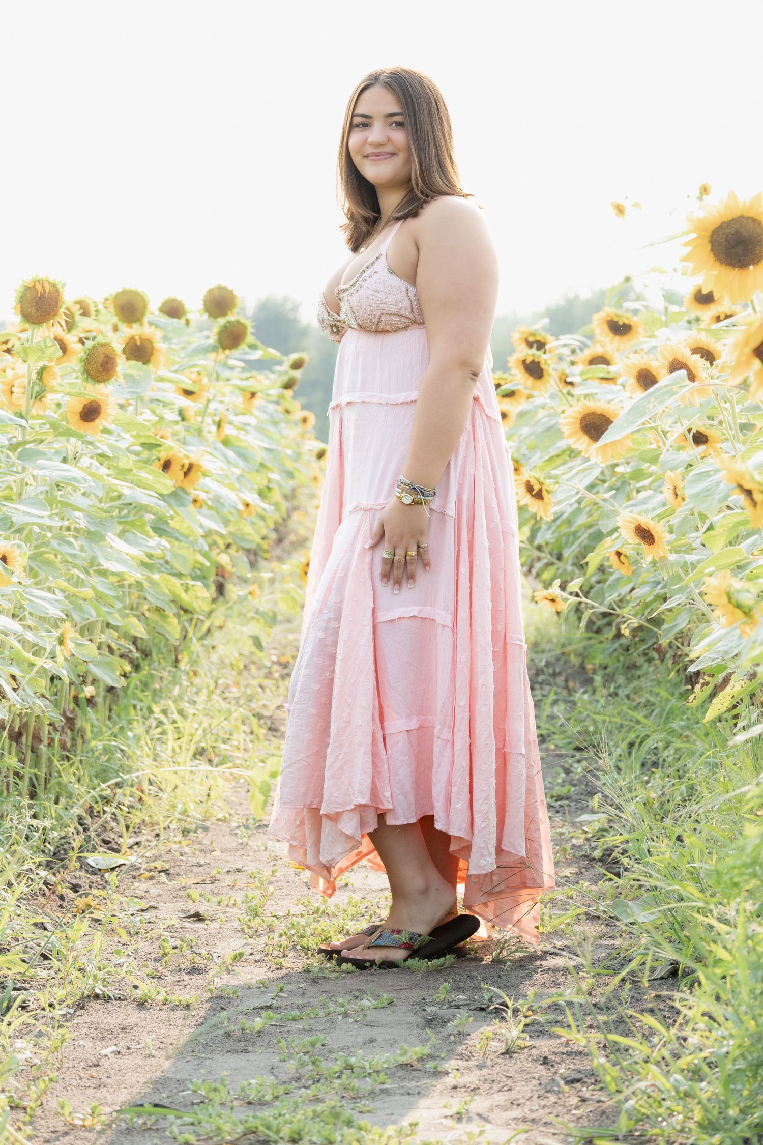 Young woman in pink boho maxi dress standing in sunflower field on sunny day.