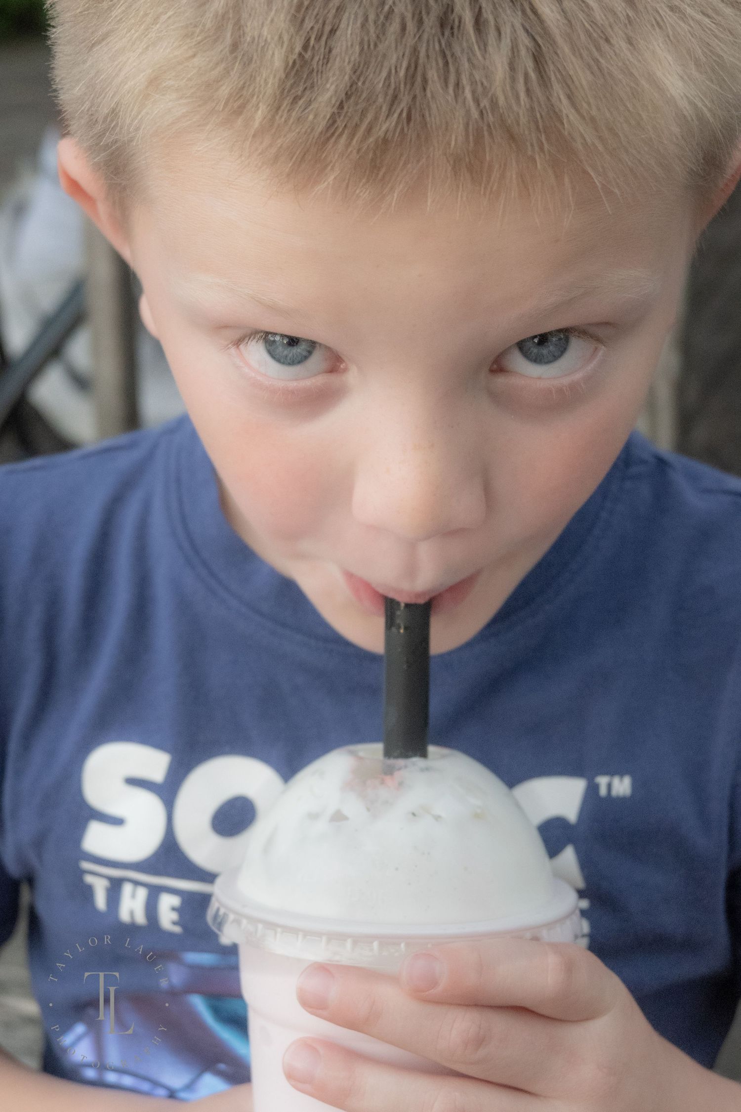 Blonde boy with blue eyes drinking a milkshake through a black straw wearing a blue Sonic t-shirt.
