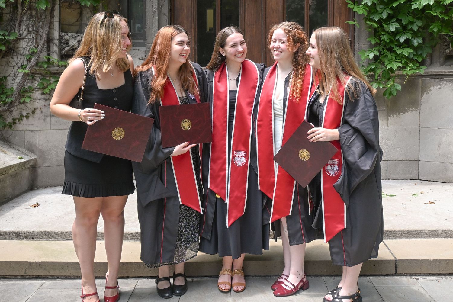 Five female college graduates in caps and gowns holding diplomas posing outdoors on campus.