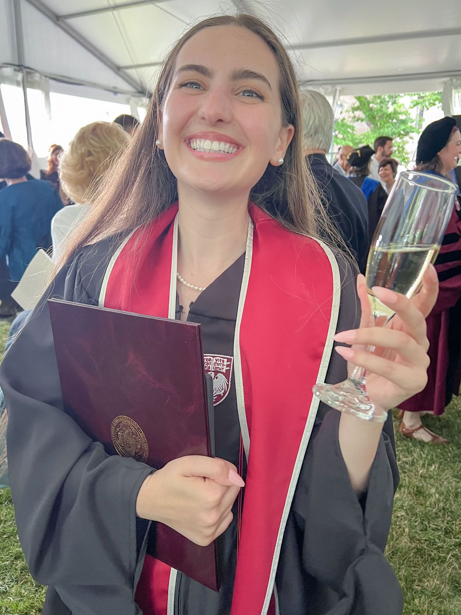 Smiling female college graduate holding diploma and champagne glass in black gown and red stole.