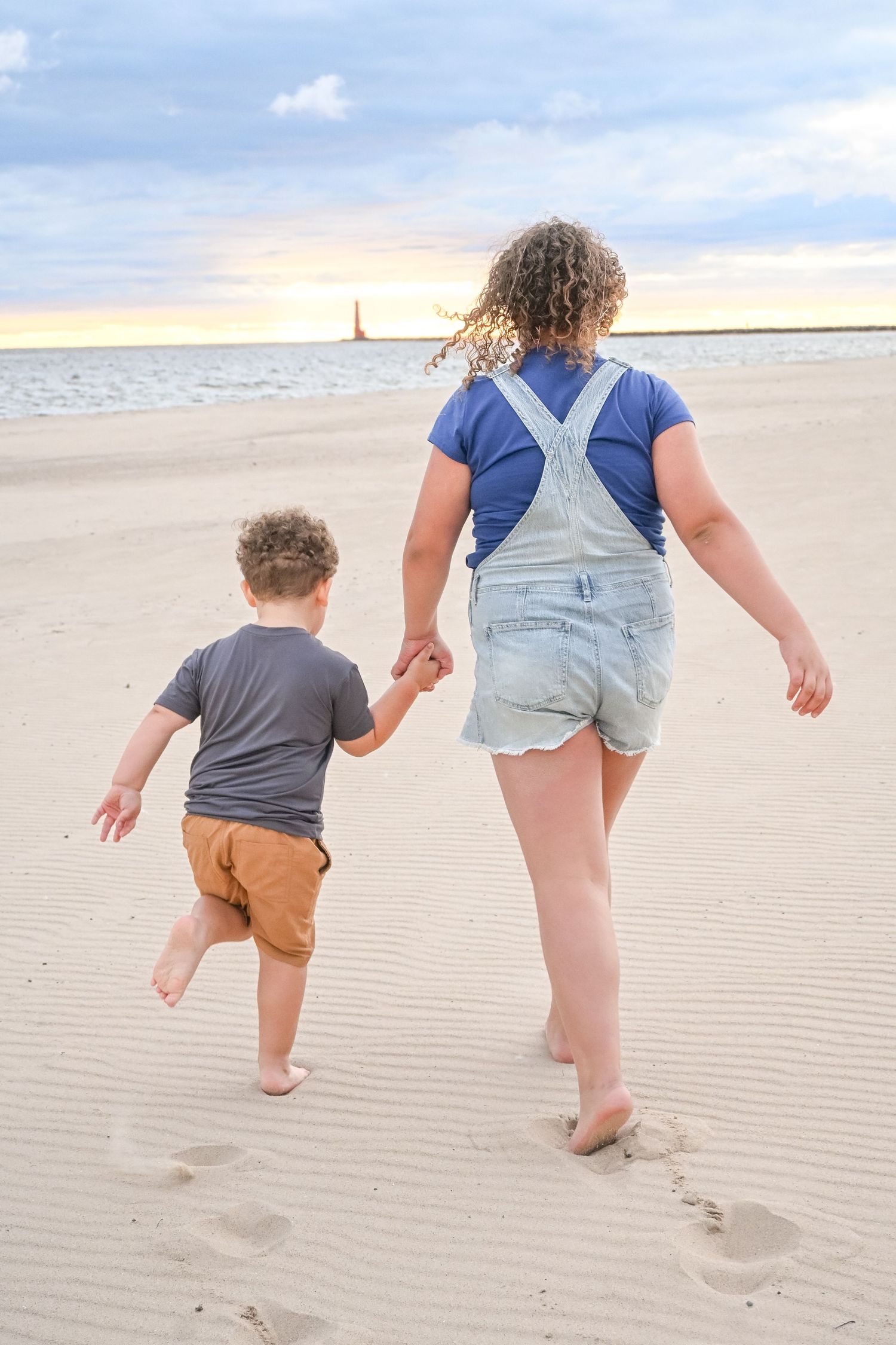 A girl in denim overalls holds hands with a young boy walking toward the ocean.
