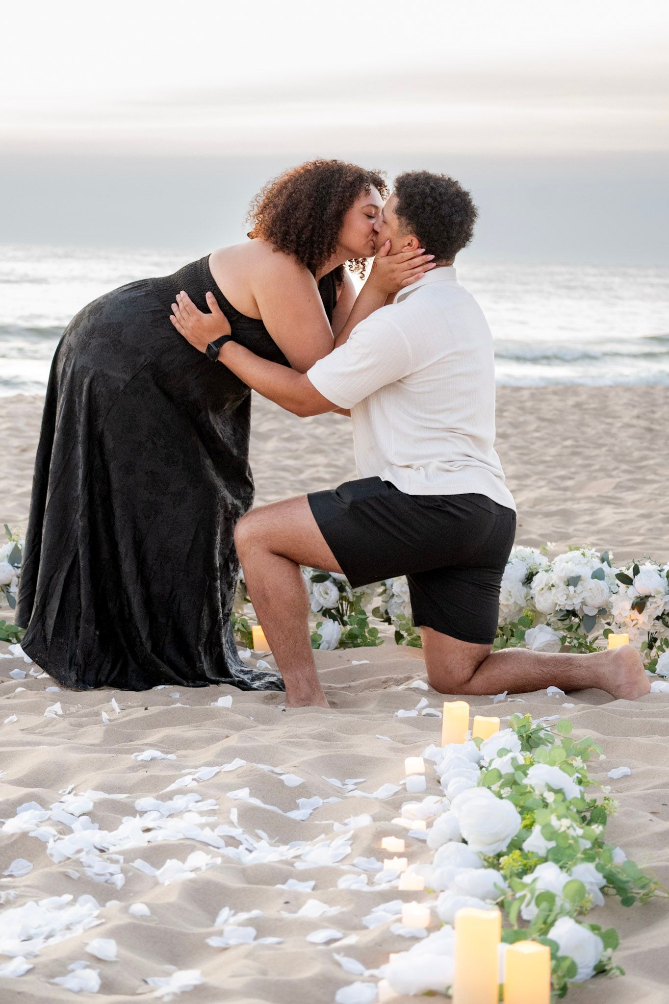 Couple sharing a romantic kiss during a beach proposal with white flowers, rose petals, and candles at sunset.