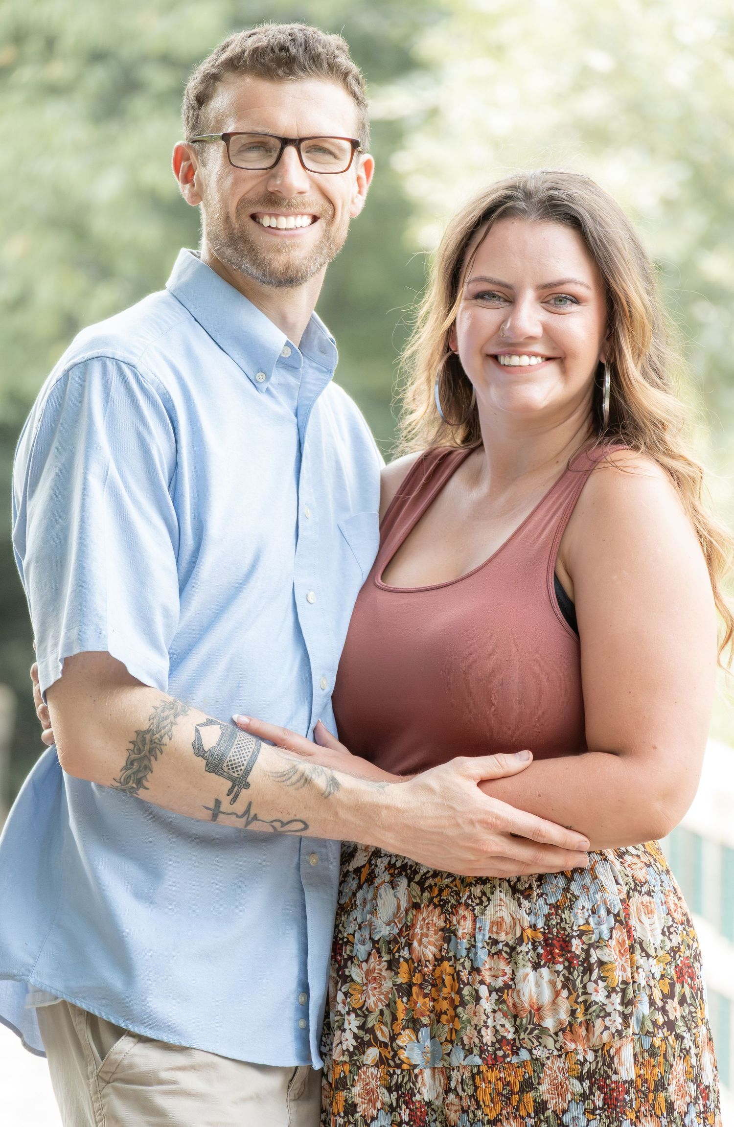 Smiling couple embracing outdoors, man in blue shirt with tattoos and glasses, woman in rust top and floral skirt.