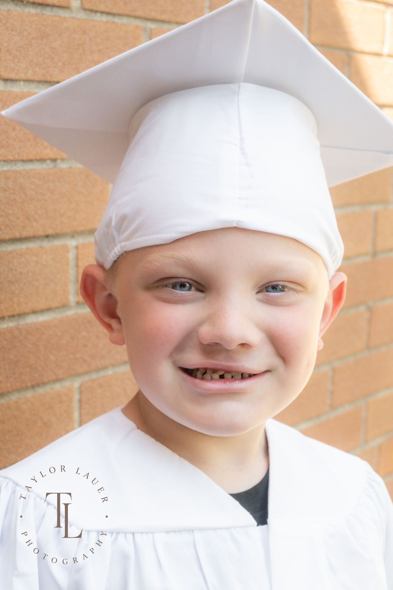 Close-up of a smiling young boy wearing a white graduation cap and gown against a brick wall.
