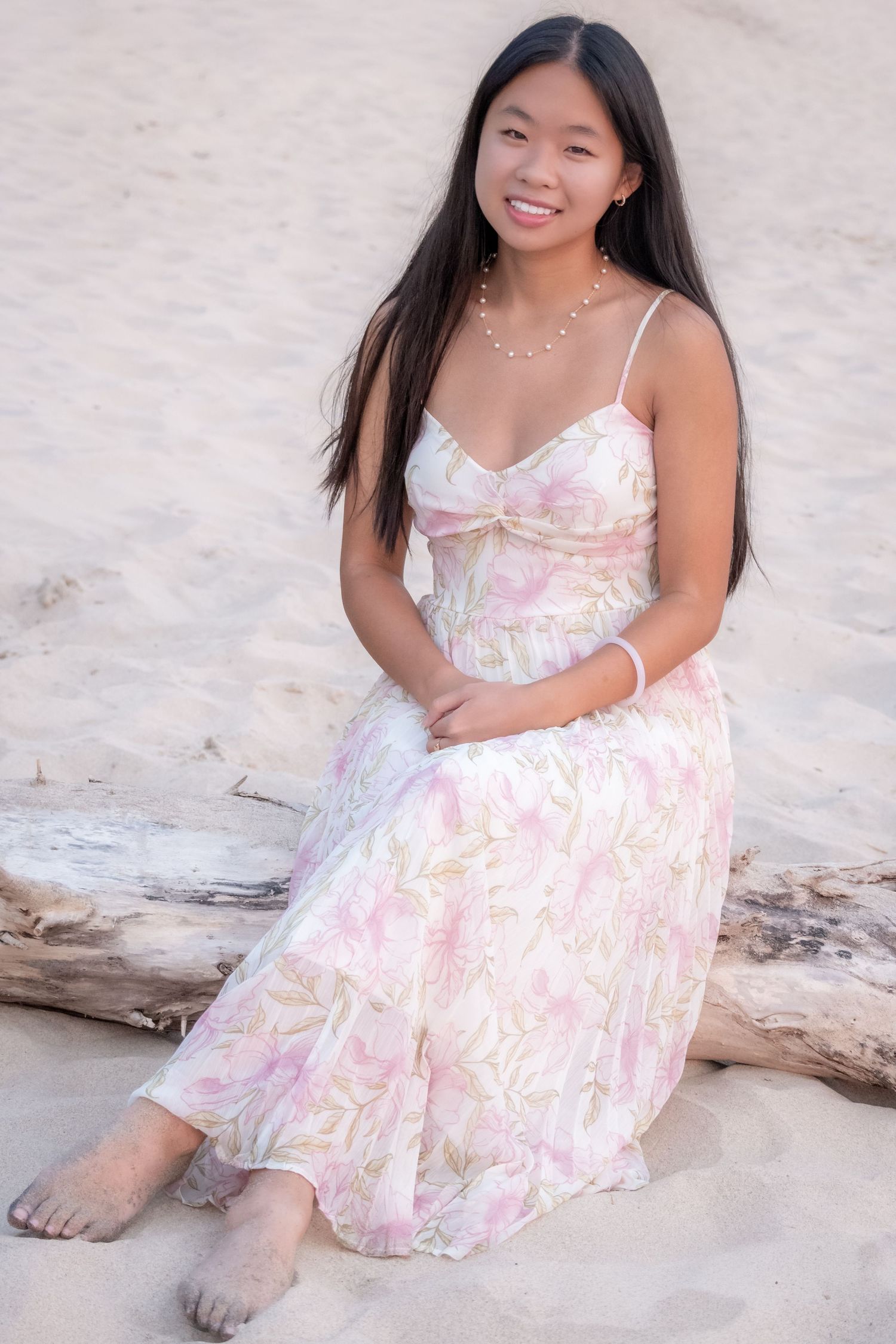 Young Asian woman in floral maxi dress sitting on driftwood at sandy beach, smiling, wearing pearl necklace.