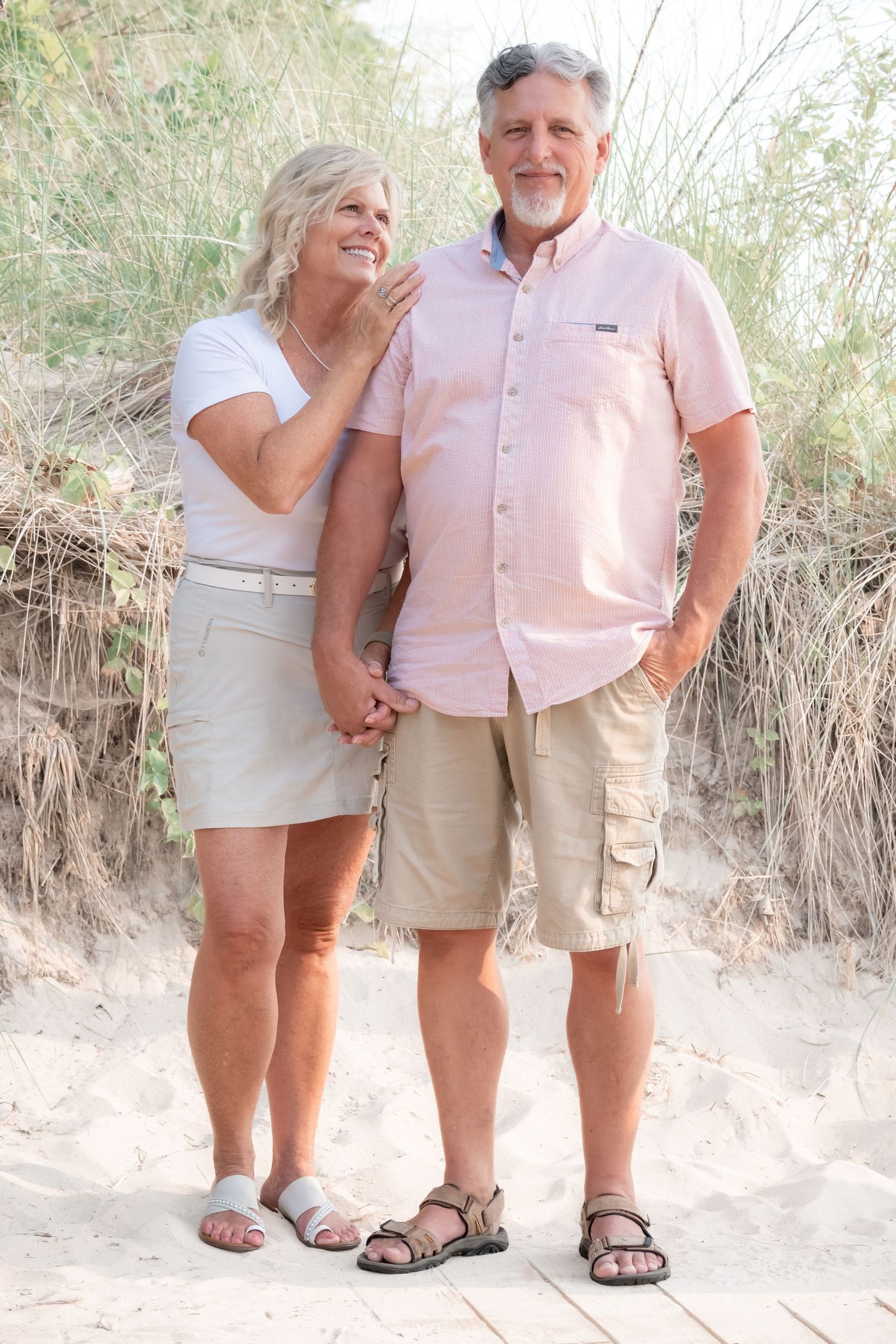 Happy senior couple holding hands on sandy beach, woman in white top and skirt, man in pink shirt and khaki shorts.