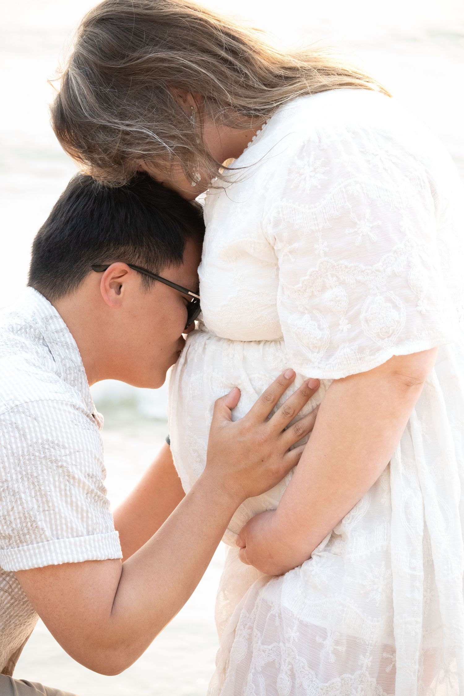 Man kneeling and kissing pregnant woman's belly while she tenderly looks down during an outdoor maternity photo session.