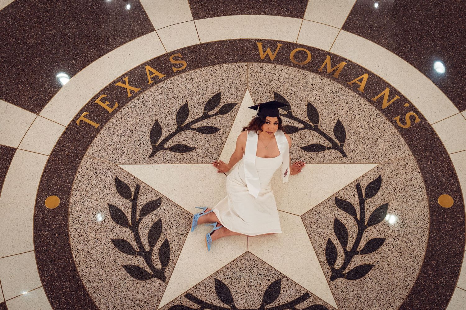 Graduate in white dress poses on Texas Woman's University star mosaic floor emblem during graduation celebration.