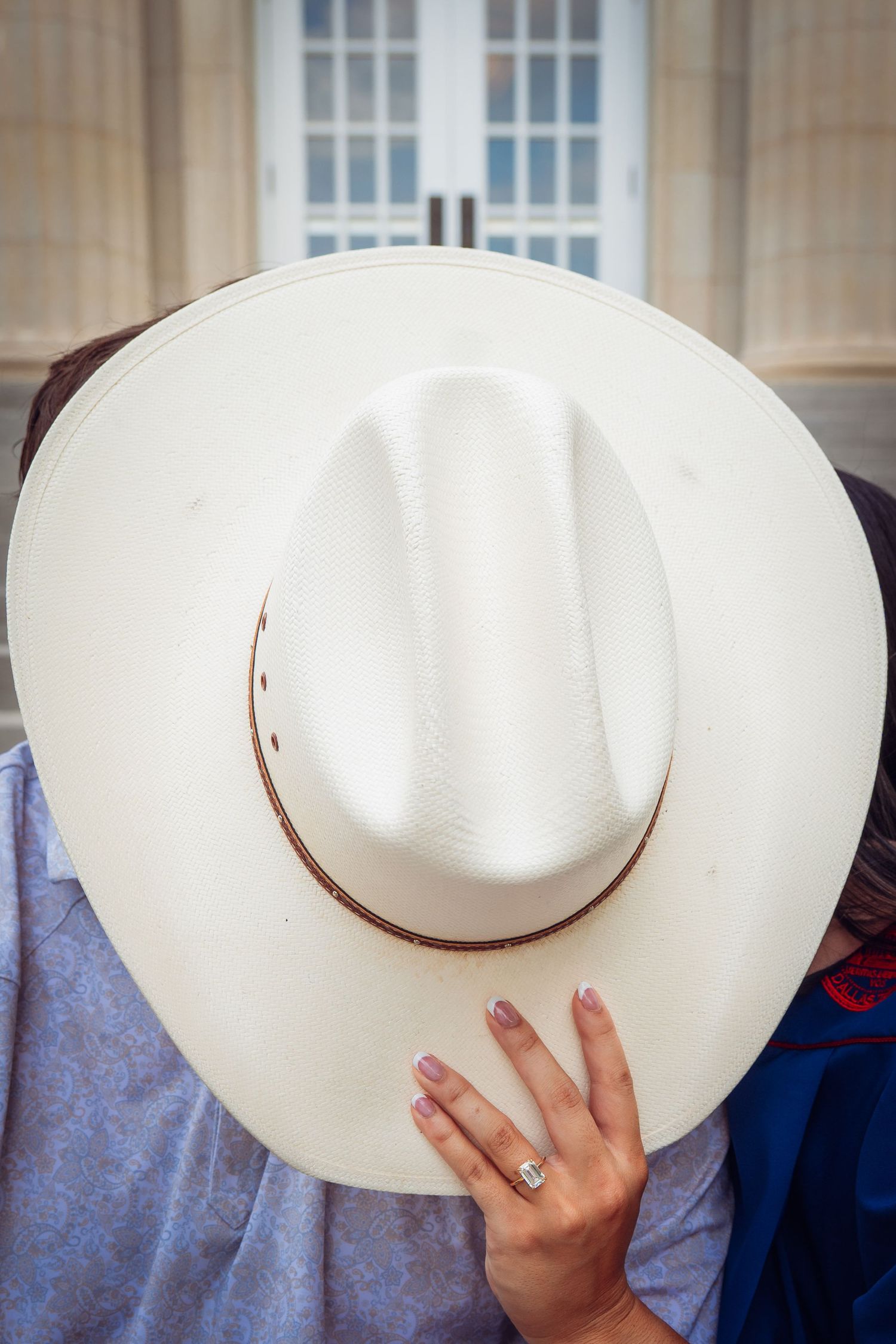 Couple sharing a kiss hidden behind a white cowboy hat, with a diamond engagement ring visible on her hand.
